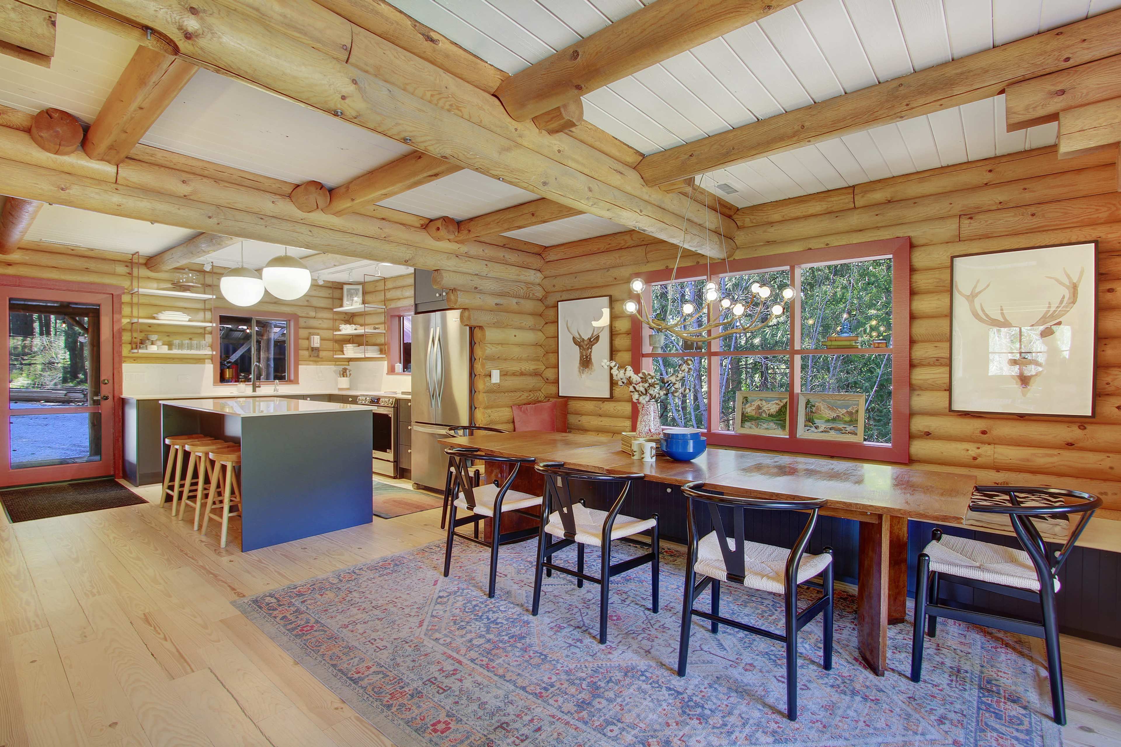 A spacious, log cabin kitchen features a central dining table, wooden beams overhead, and large windows overlooking a natural setting.