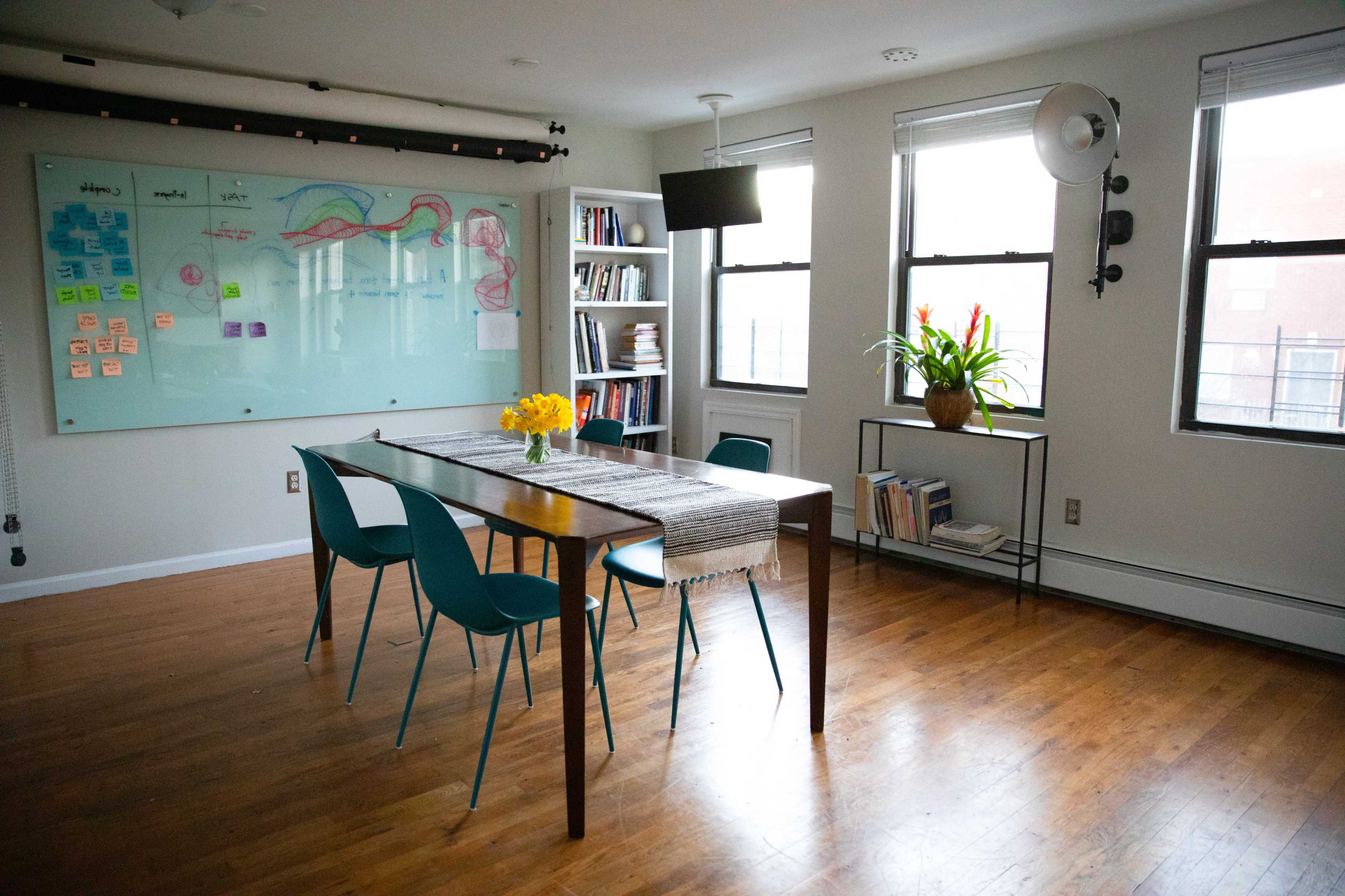 A bright, organized room with a wooden table surrounded by teal chairs, a whiteboard covered in notes, and shelves filled with books.