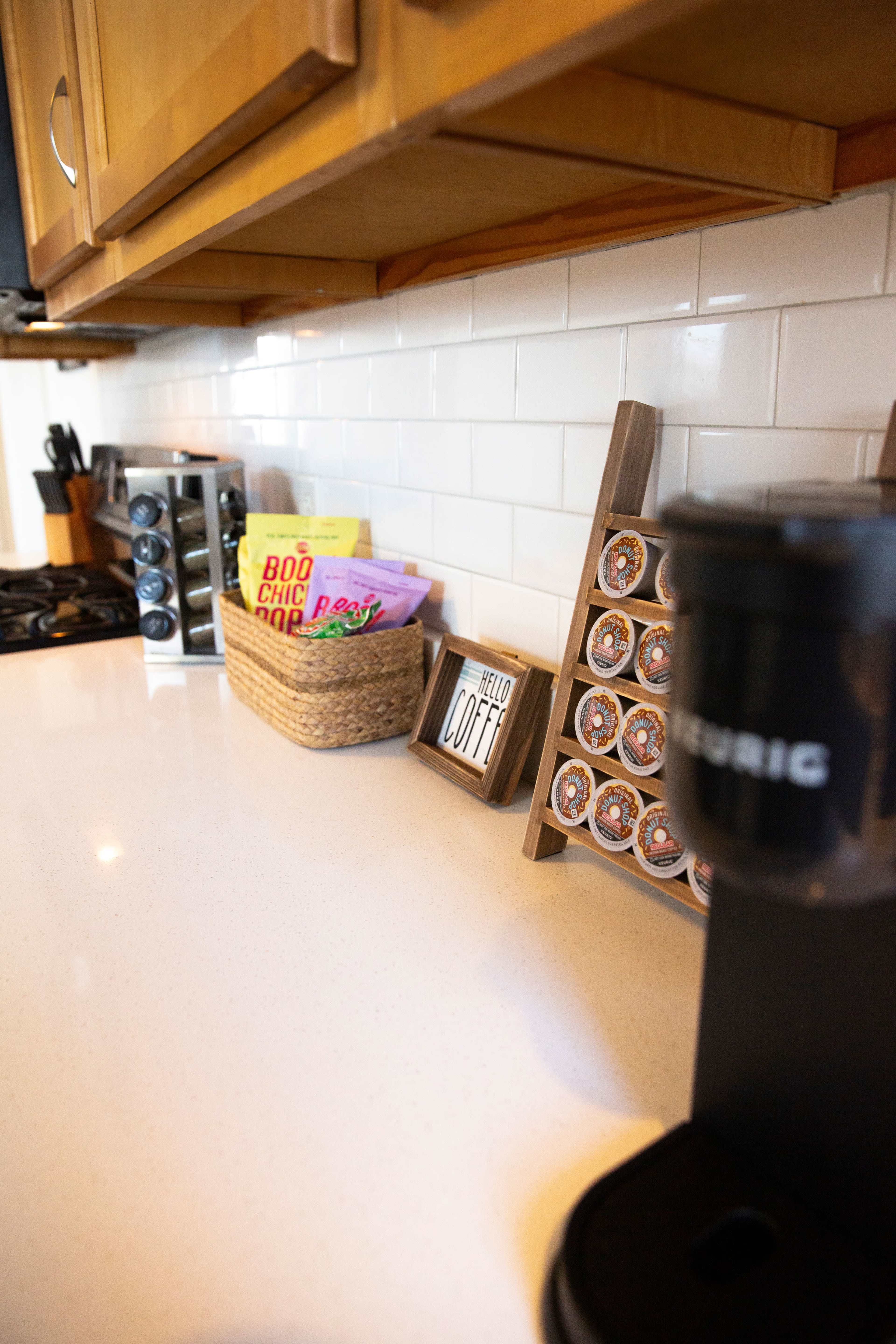 A kitchen countertop displays a coffee machine, a rack of coffee pods, and a woven basket containing snacks.