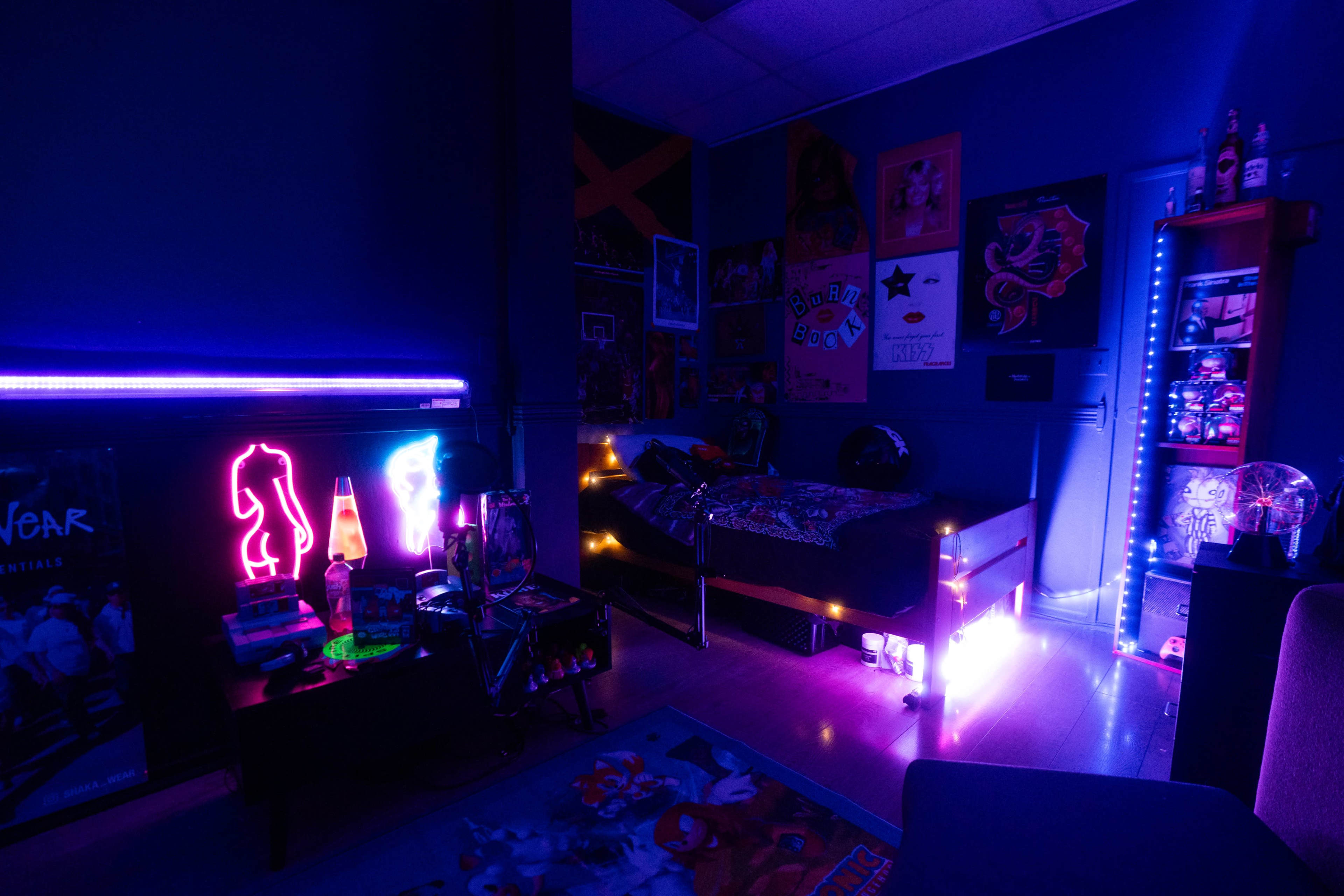 The image shows a dimly lit bedroom featuring a bed, colorful neon lights, posters on the walls, and a shelf with various items.