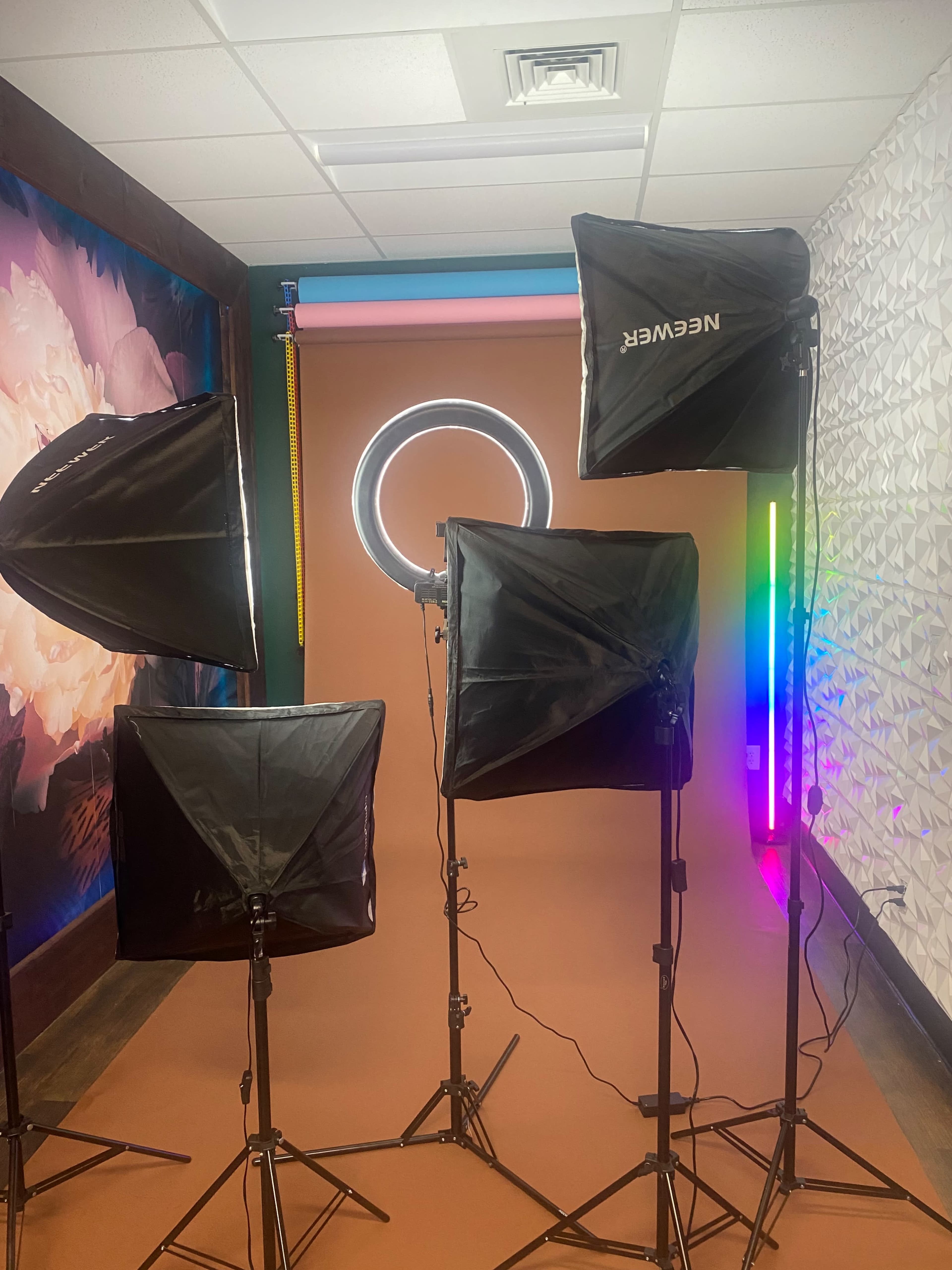 The image shows a photography studio setup featuring multiple softbox lights and a circular LED light against a backdrop with colorful lighting and a textured wall.