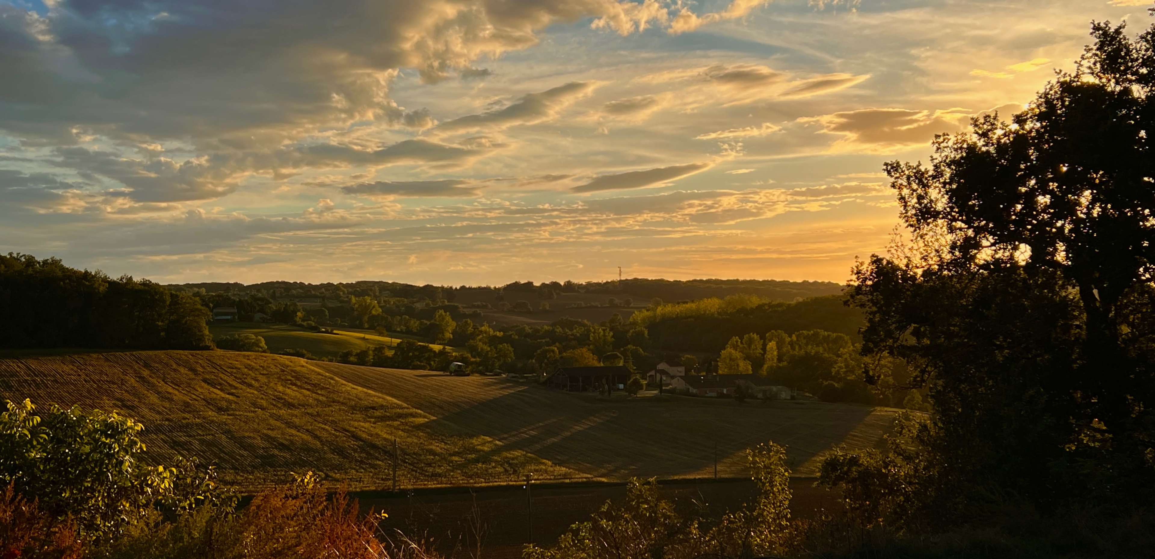 The scene features rolling hills and fields under a colorful sky at sunset with scattered clouds and a small cluster of houses in the valley.