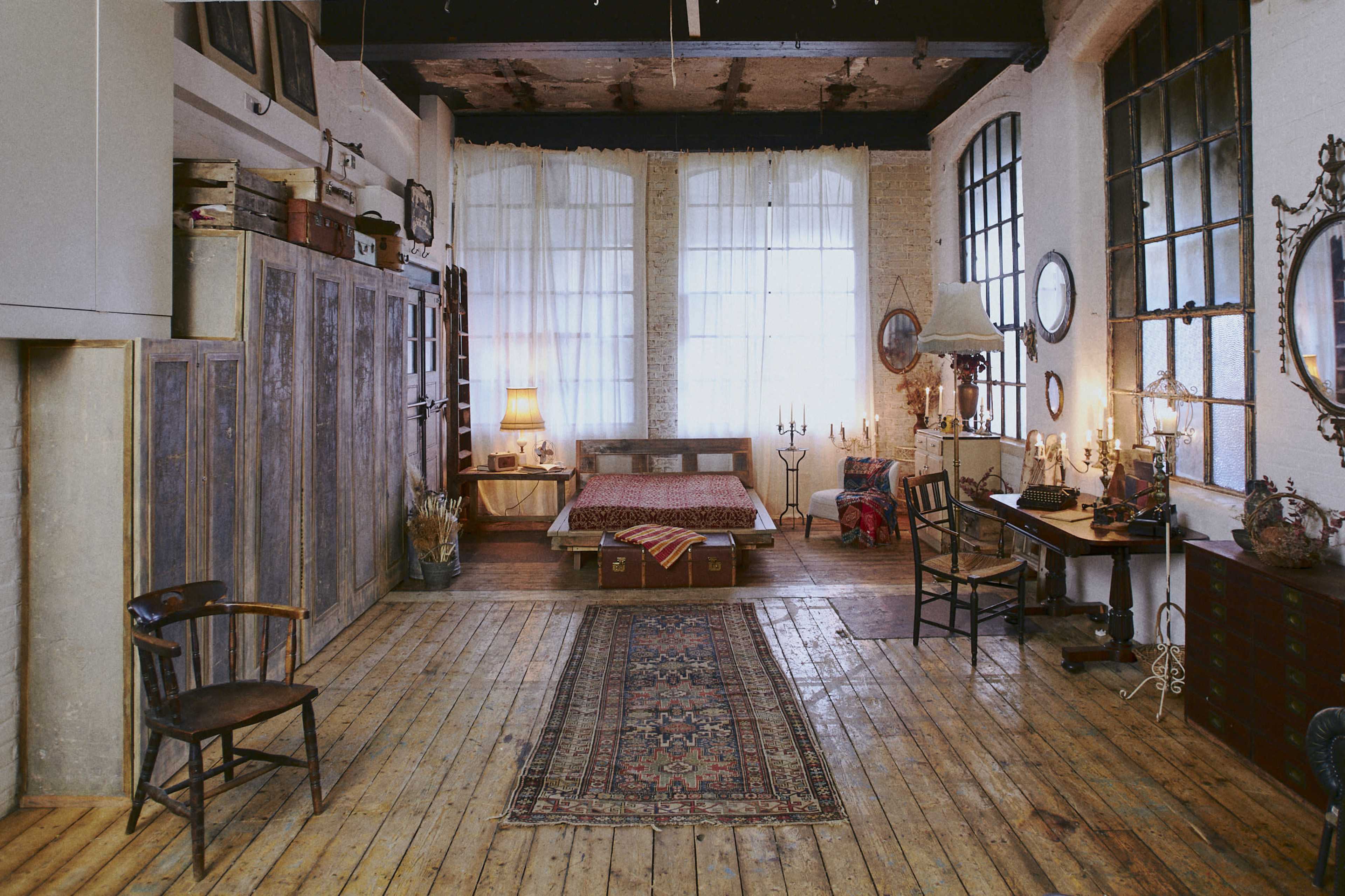 A spacious, rustic bedroom with large windows, wooden floors, a bed in the center, and various pieces of furniture including a chair, desk, and wardrobe.