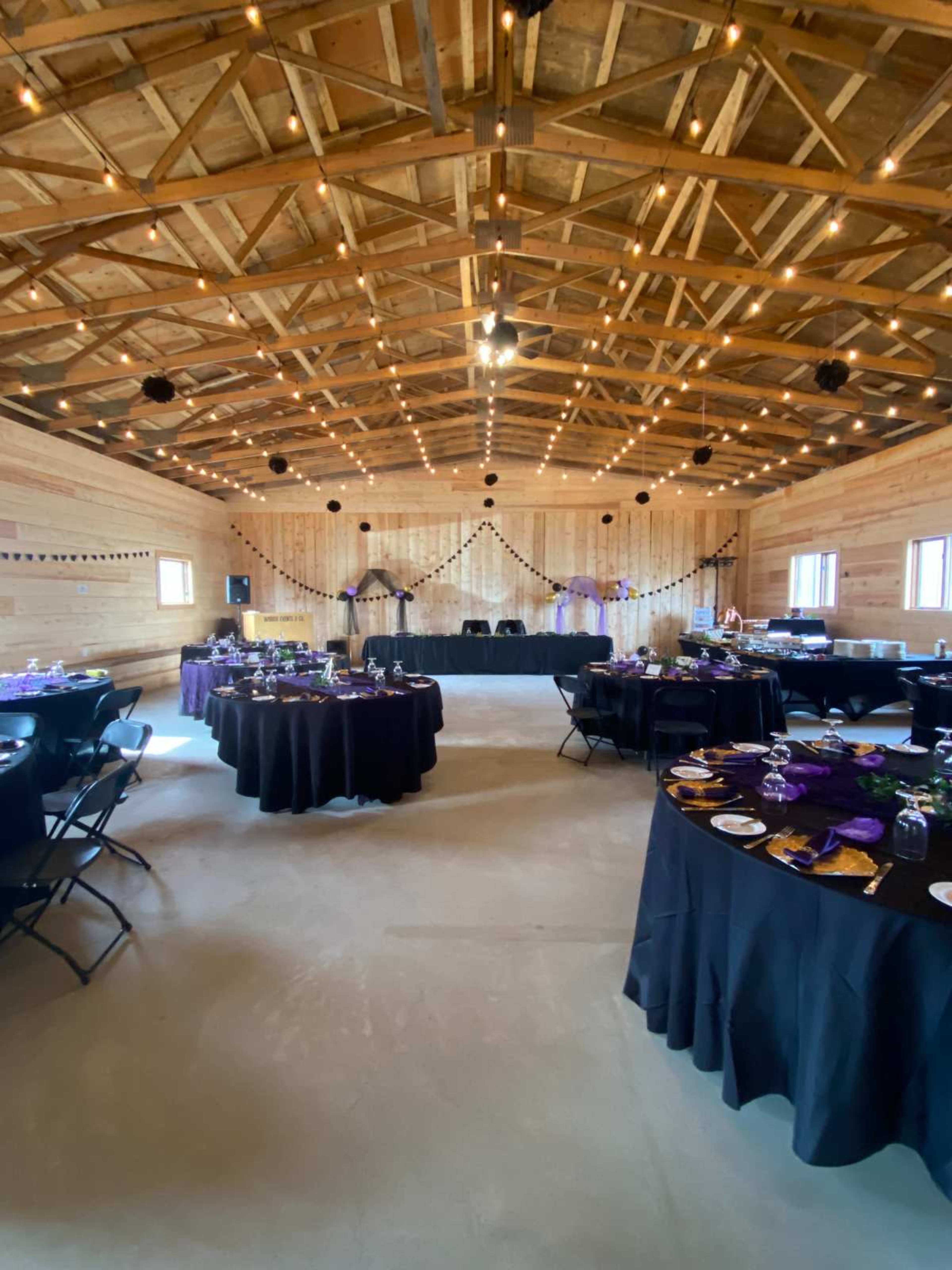 The image shows a decorated event space with black tables set for a gathering, featuring wooden walls and overhead string lights.