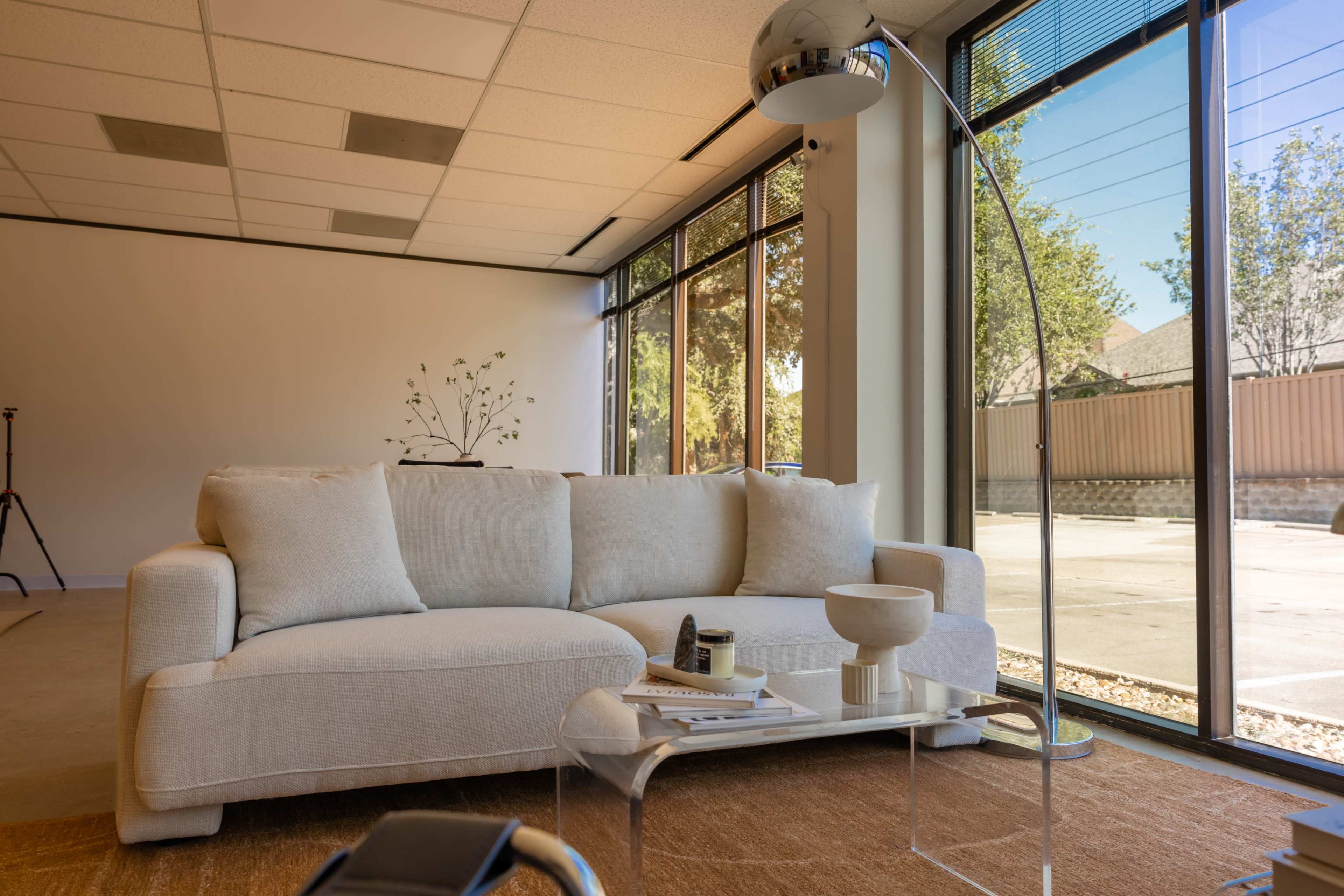 The image shows a modern living room with a light-colored sofa, a glass coffee table, and large windows allowing natural light to fill the space.