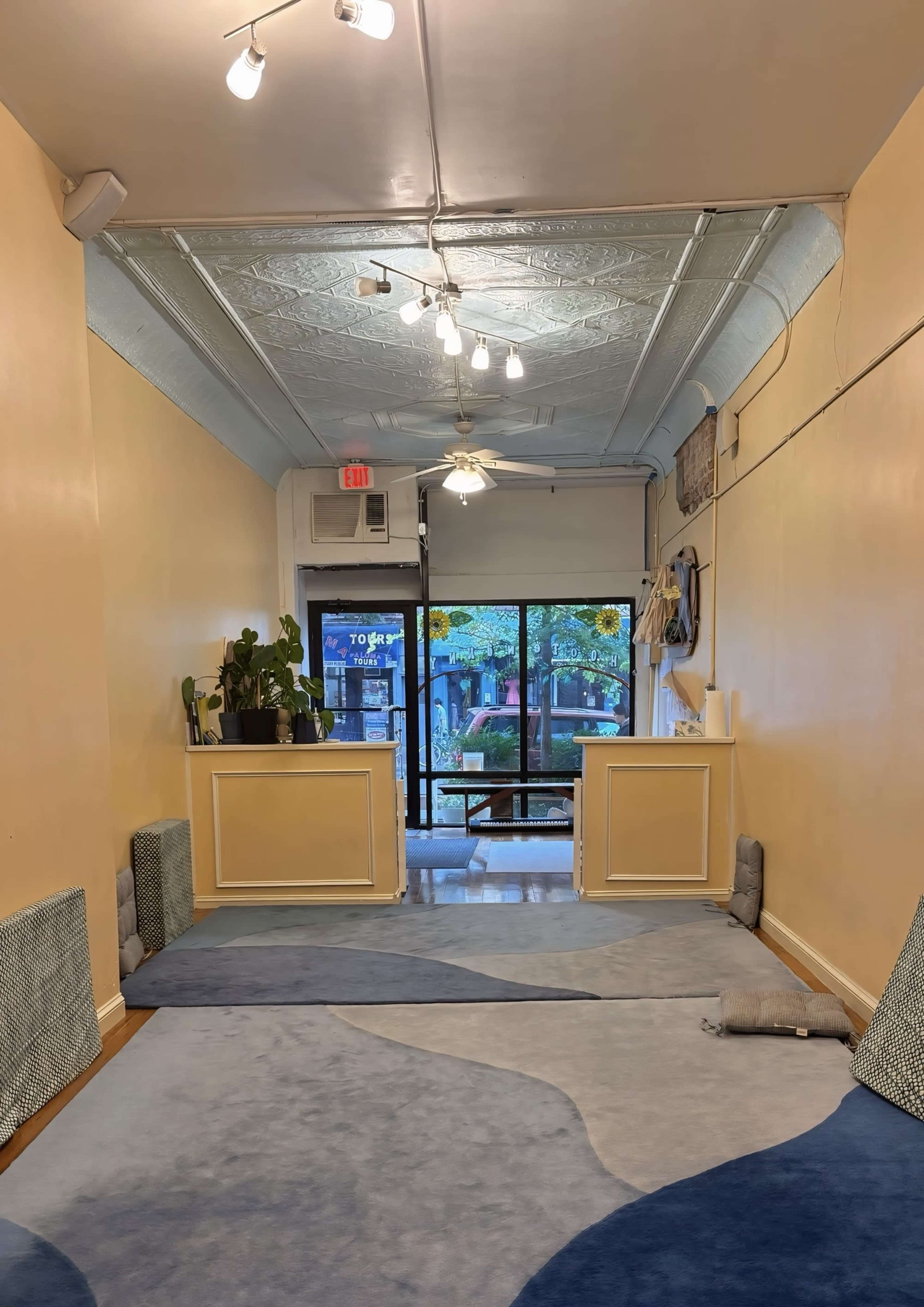 The image shows a narrow hallway with light-colored walls, decorative rugs on the floor, and glass doors at the end leading to an outdoor area.