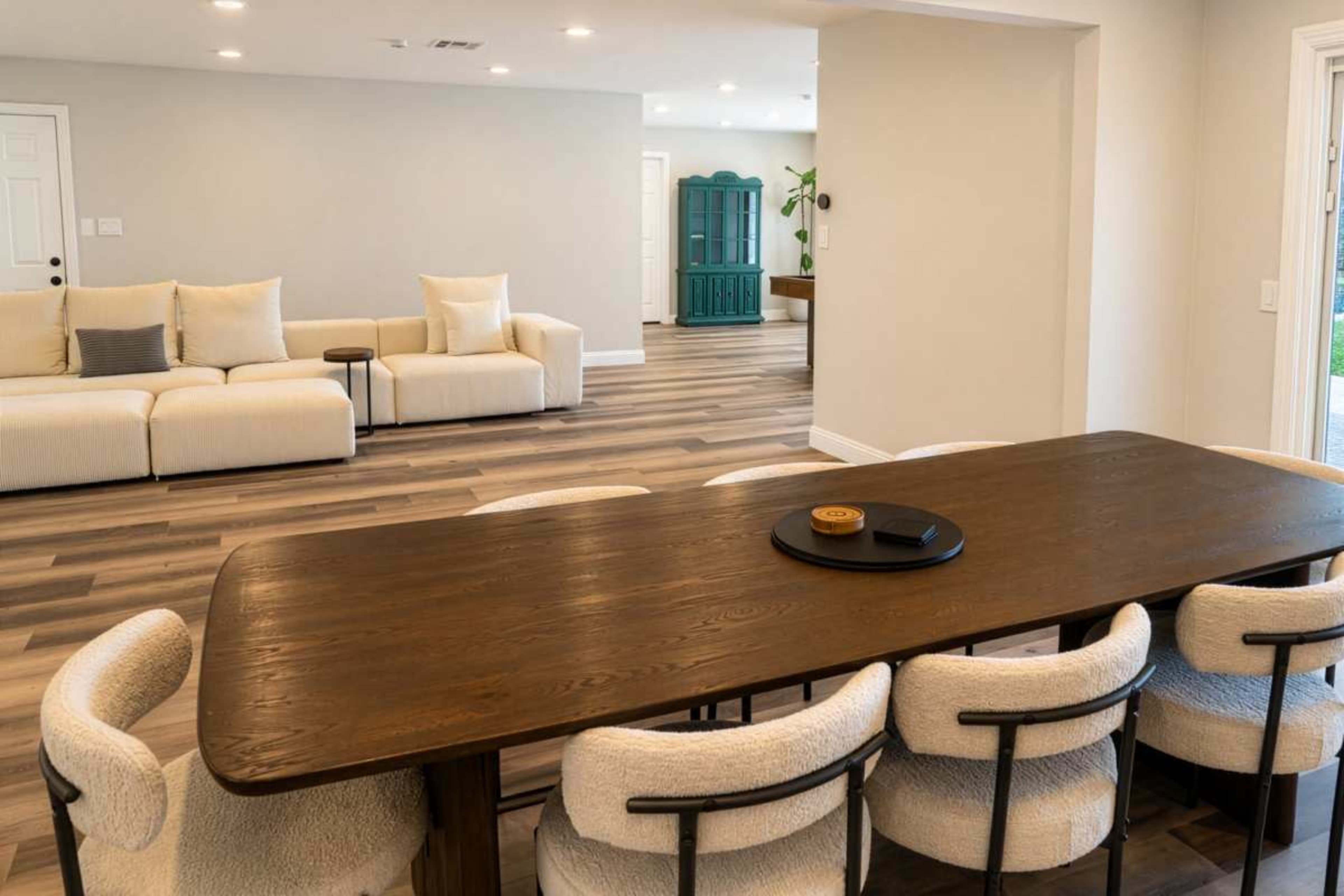 The image shows a modern dining area with a long wooden table surrounded by upholstered chairs, and a light-colored sofa visible in the background.