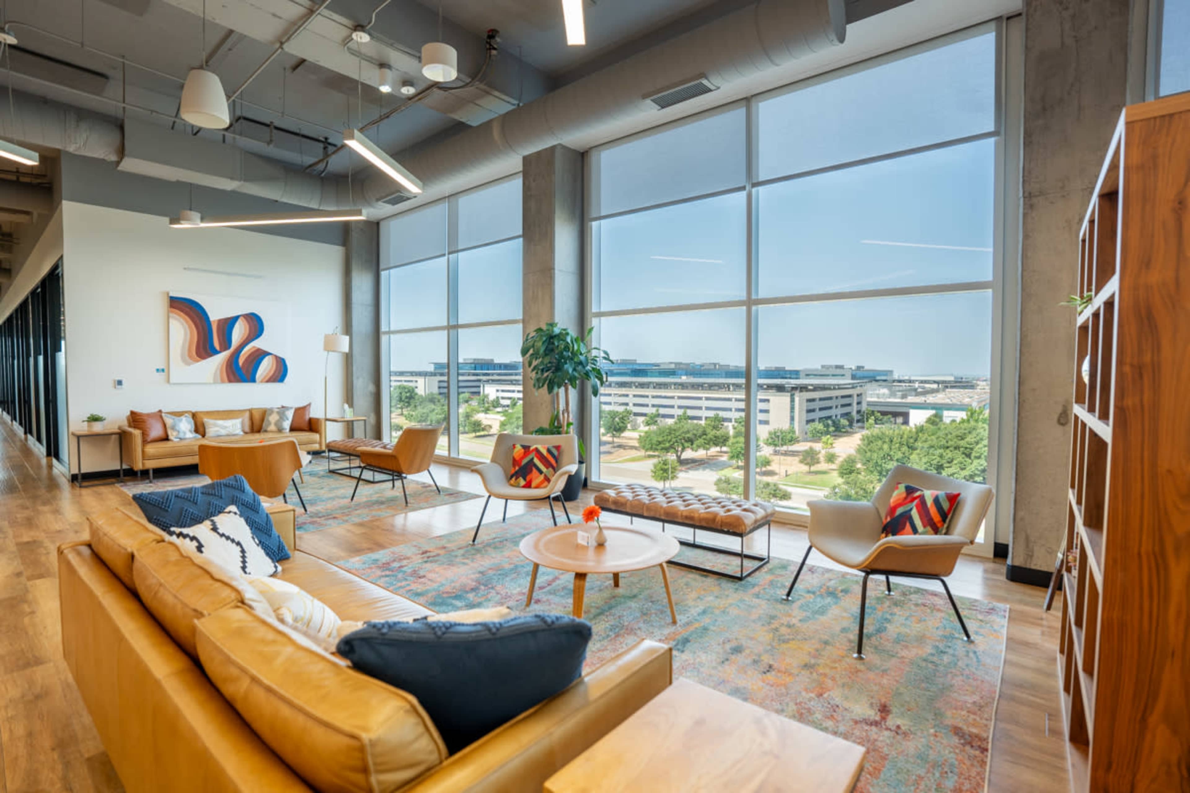 The image shows a modern office lounge area featuring a sofa, chairs, and large windows overlooking a landscaped outdoor space.