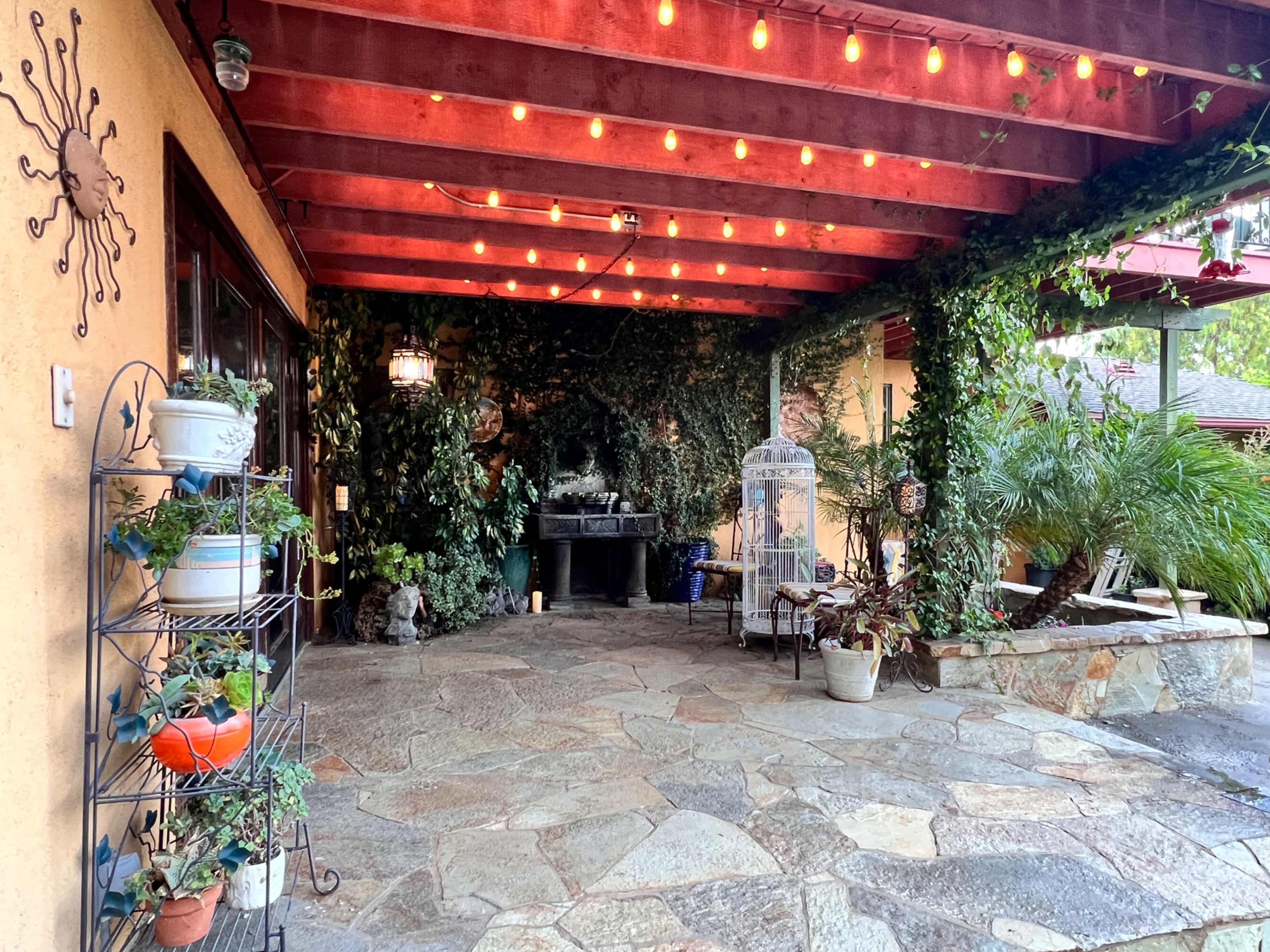 The image shows a covered outdoor patio with stone flooring, adorned with hanging lights and surrounded by greenery.