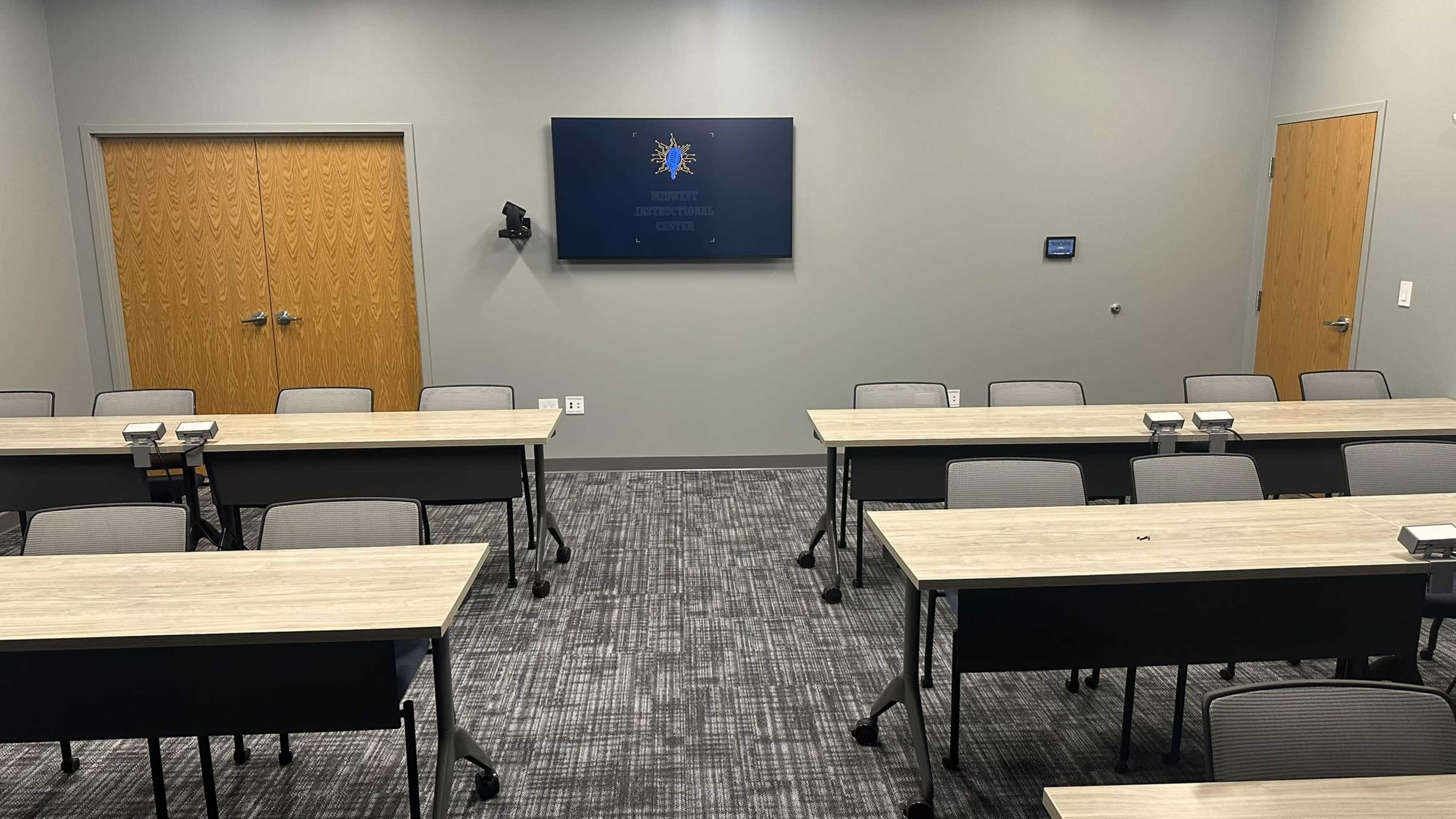 The image shows a conference room with several rows of desks facing a wall-mounted screen, and two wooden doors on the side.