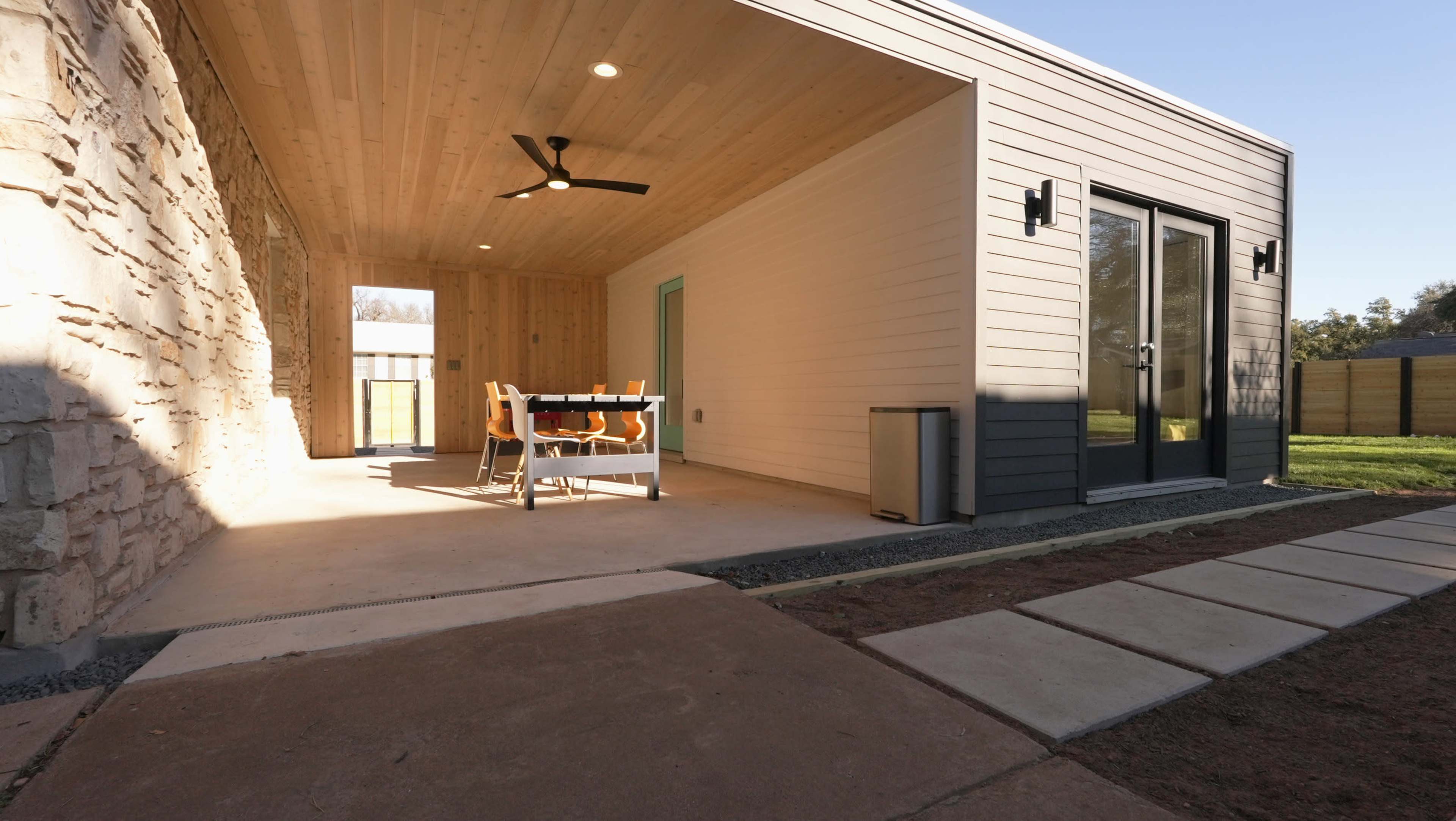 The image shows a modern outdoor patio area with a dining table and chairs, featuring a concrete pathway and a wooden ceiling.