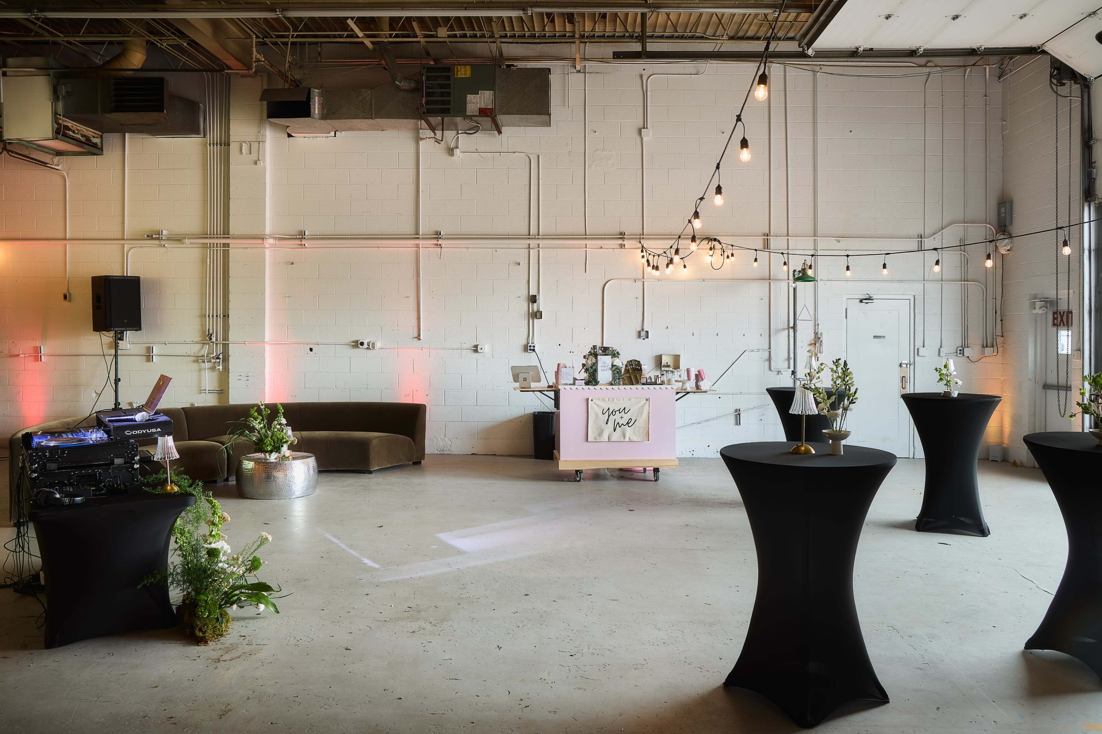 An indoor event space features a bar setup, several black cocktail tables, a DJ station, and decorative lighting.