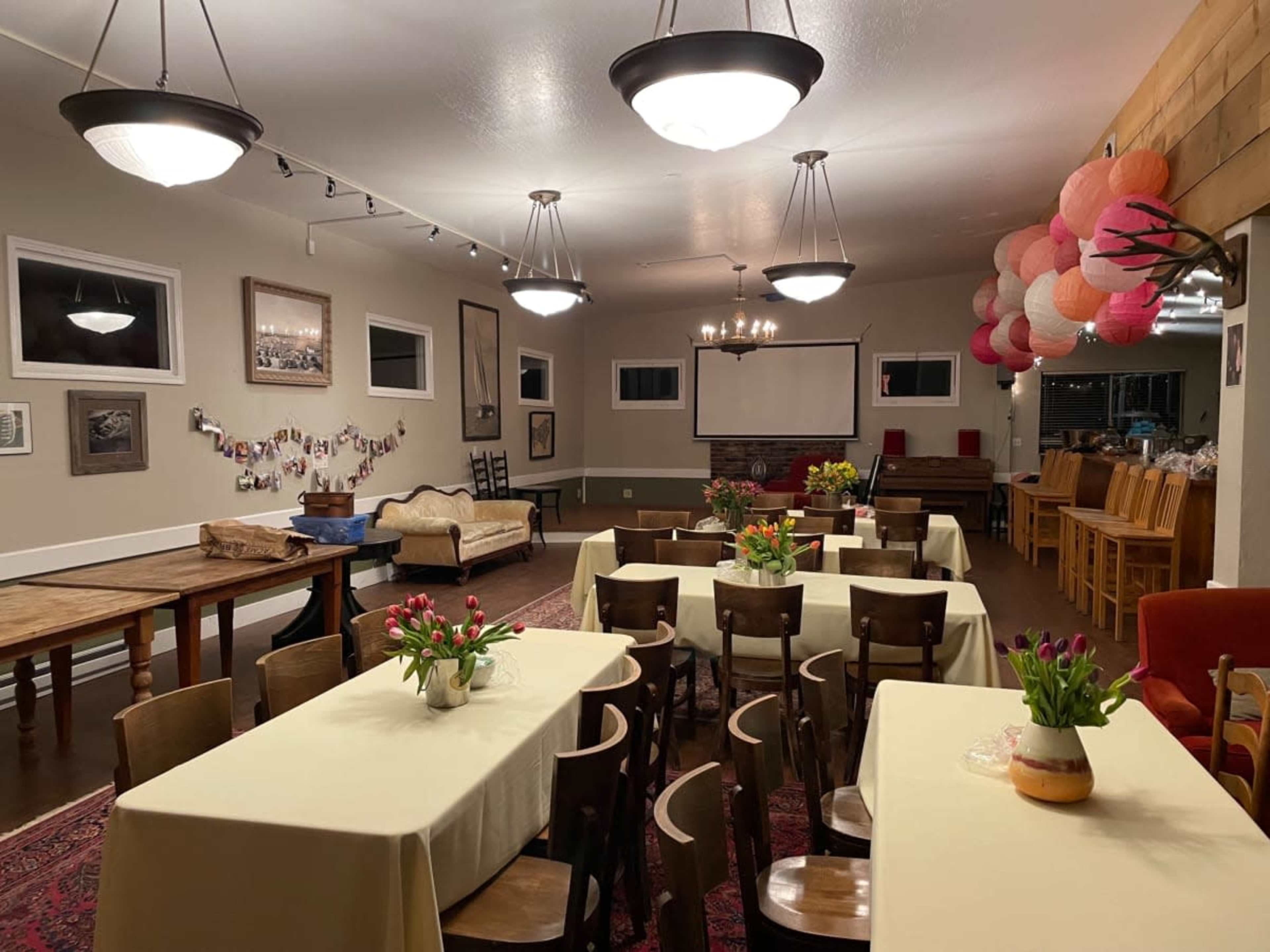 The interior of a brightly lit room features several tables with light-colored tablecloths, wooden chairs, and decorations like flowers and lanterns hanging from the ceiling.