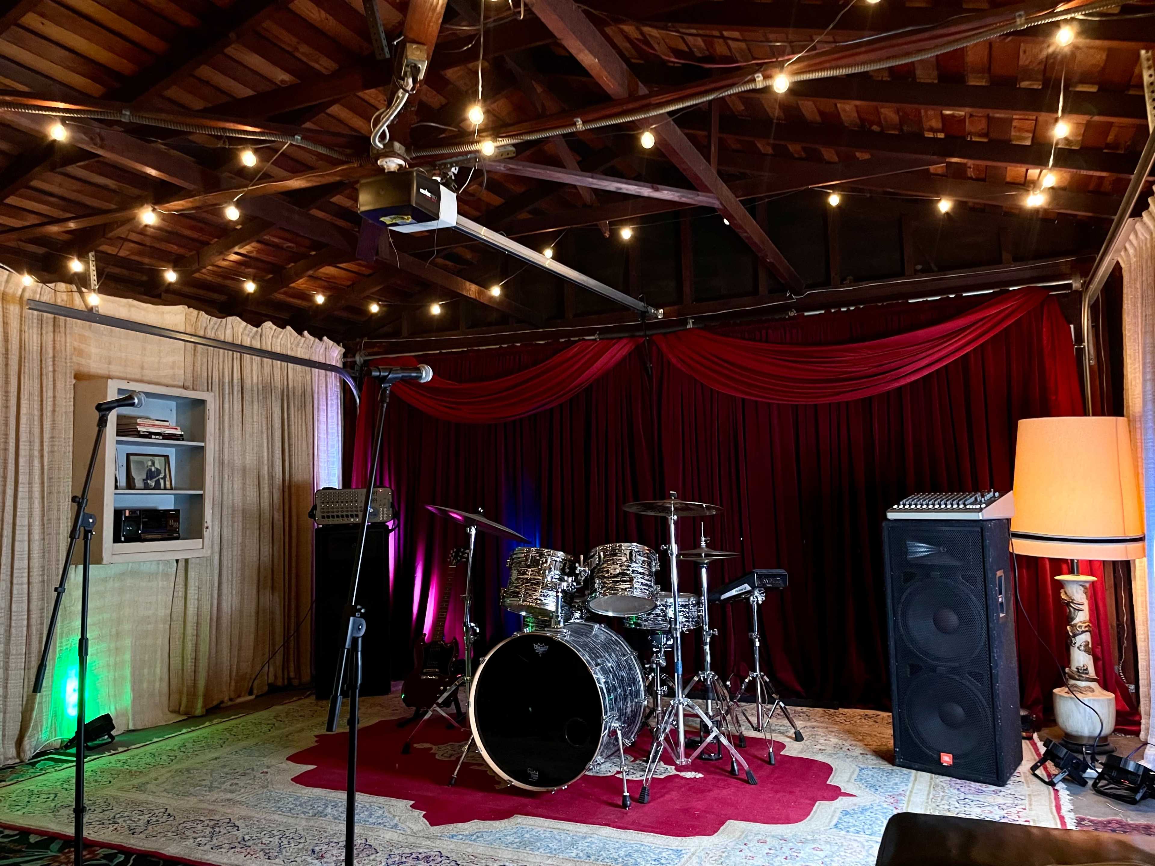 A music rehearsal space with a drum set, amplifiers, a microphone stand, and soft lighting from hanging lights and a lamp.