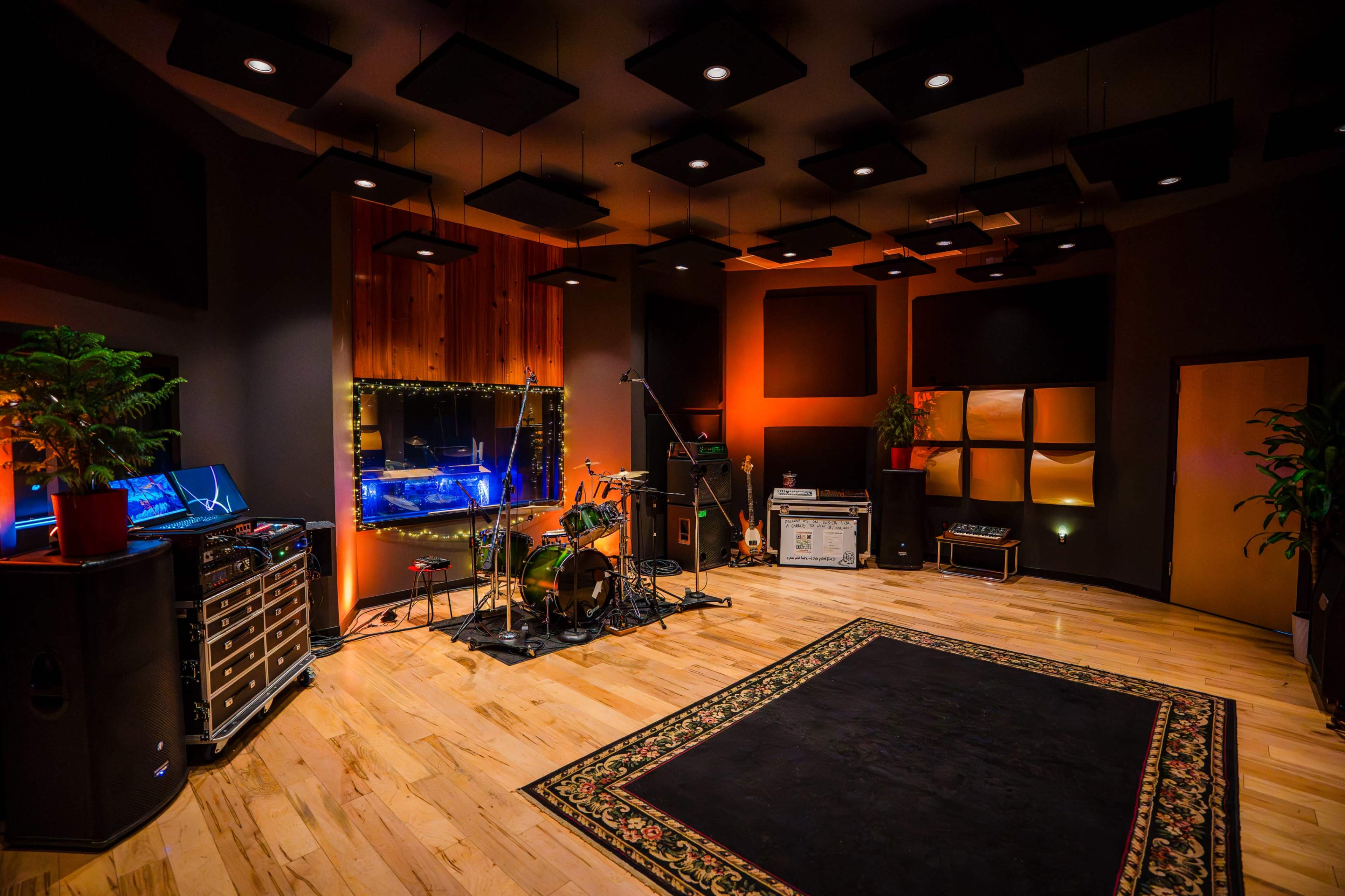 The image shows a music studio featuring wooden floors, a drum set, amplifiers, and soundproofing panels on the walls.