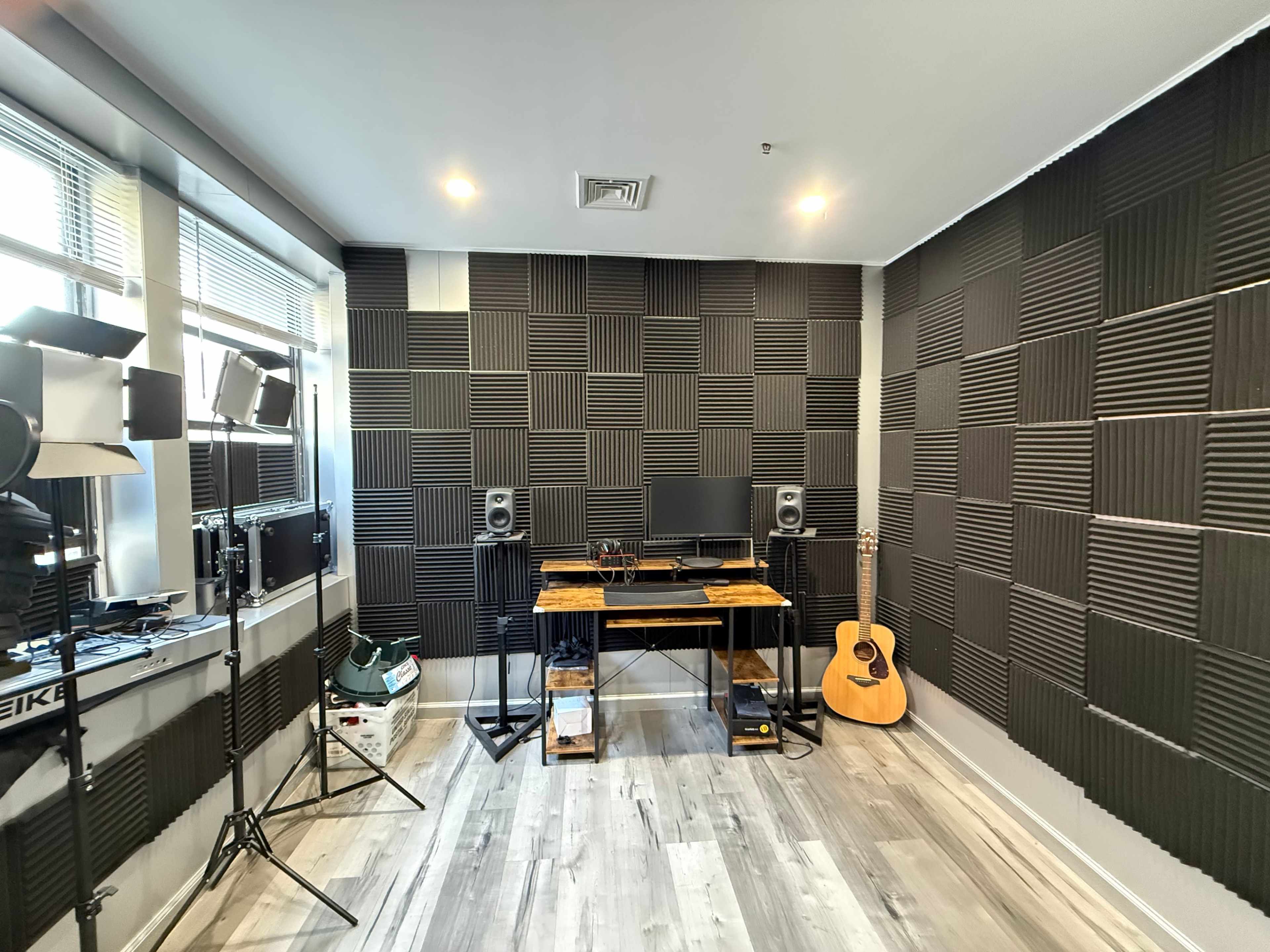 The image shows a soundproofed recording studio with a wooden desk, computer monitors, speakers, and an acoustic guitar placed in the corner.