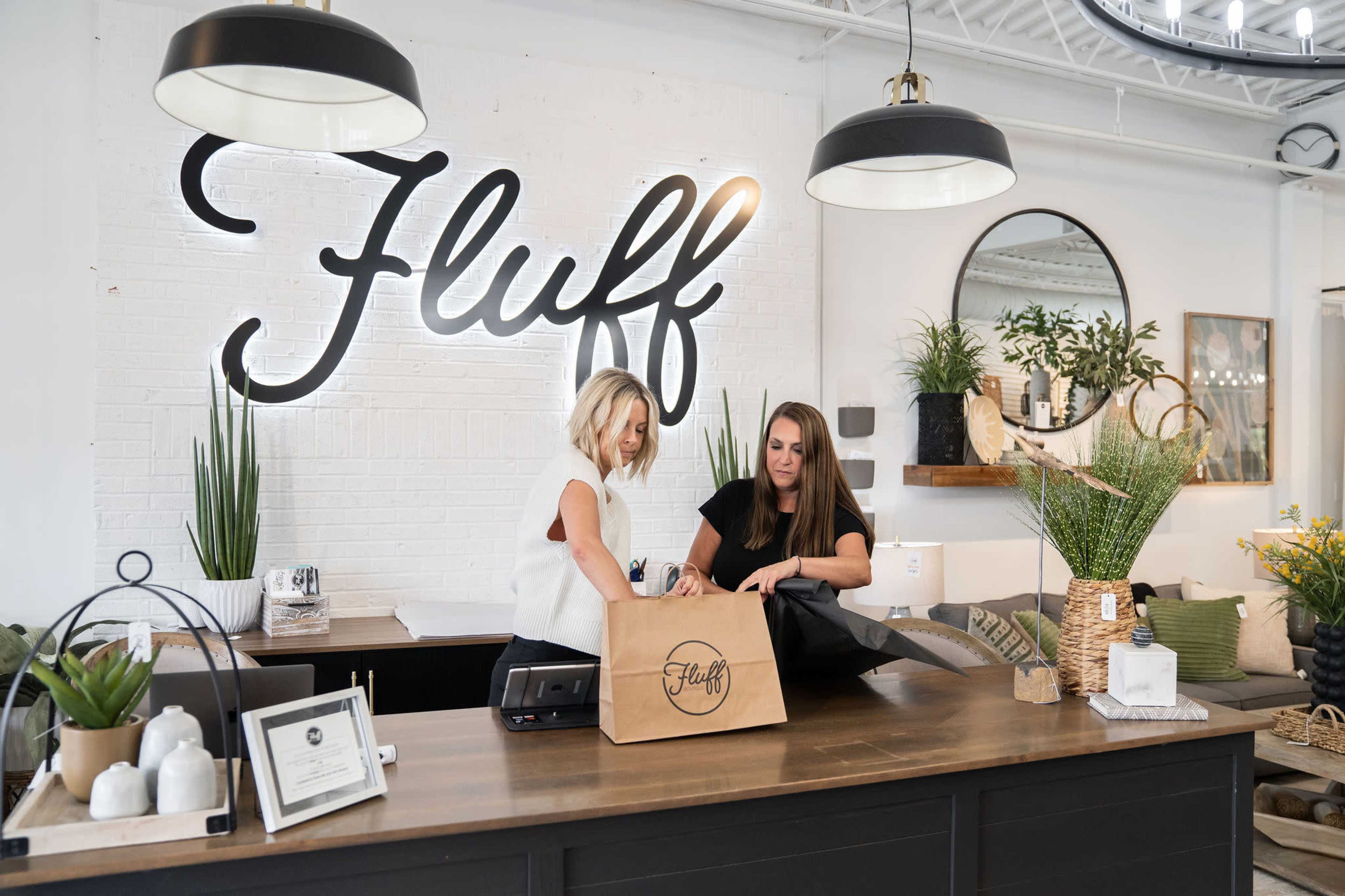 Two women are standing behind a wooden counter in a店 with a large sign that reads "Fluff," discussing a paper bag, surrounded by decorative plants and home goods.