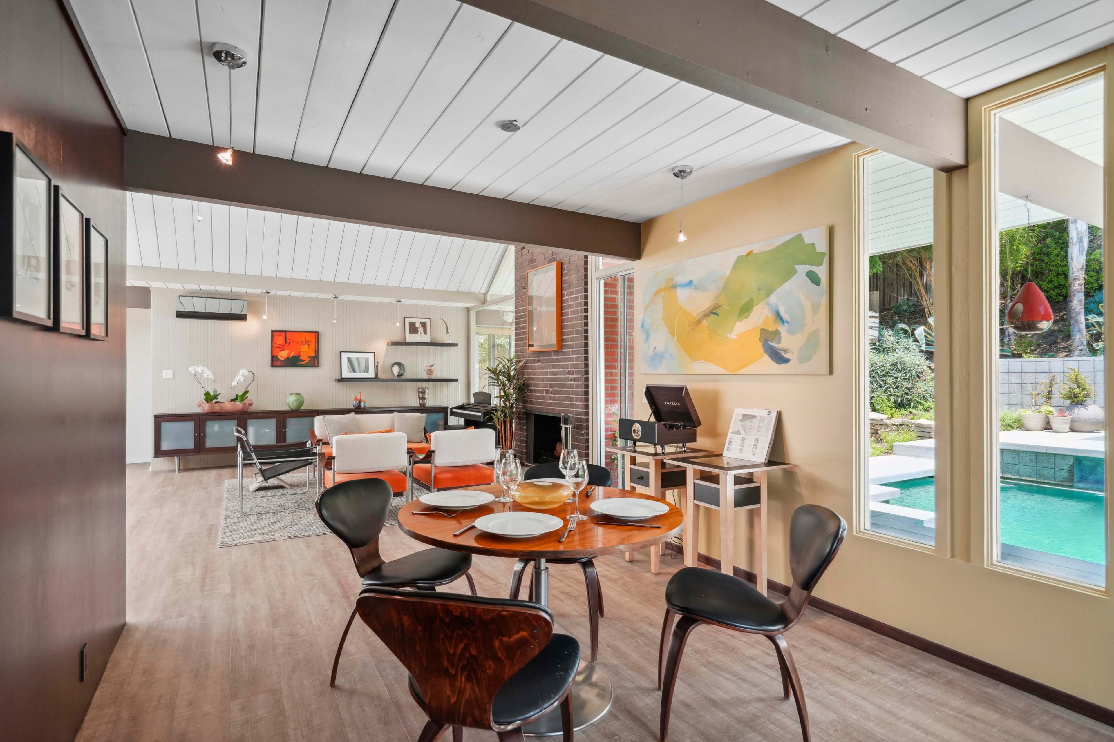 A modern dining area features a round table with black chairs, leading to a spacious living room with large windows and a view of a pool.