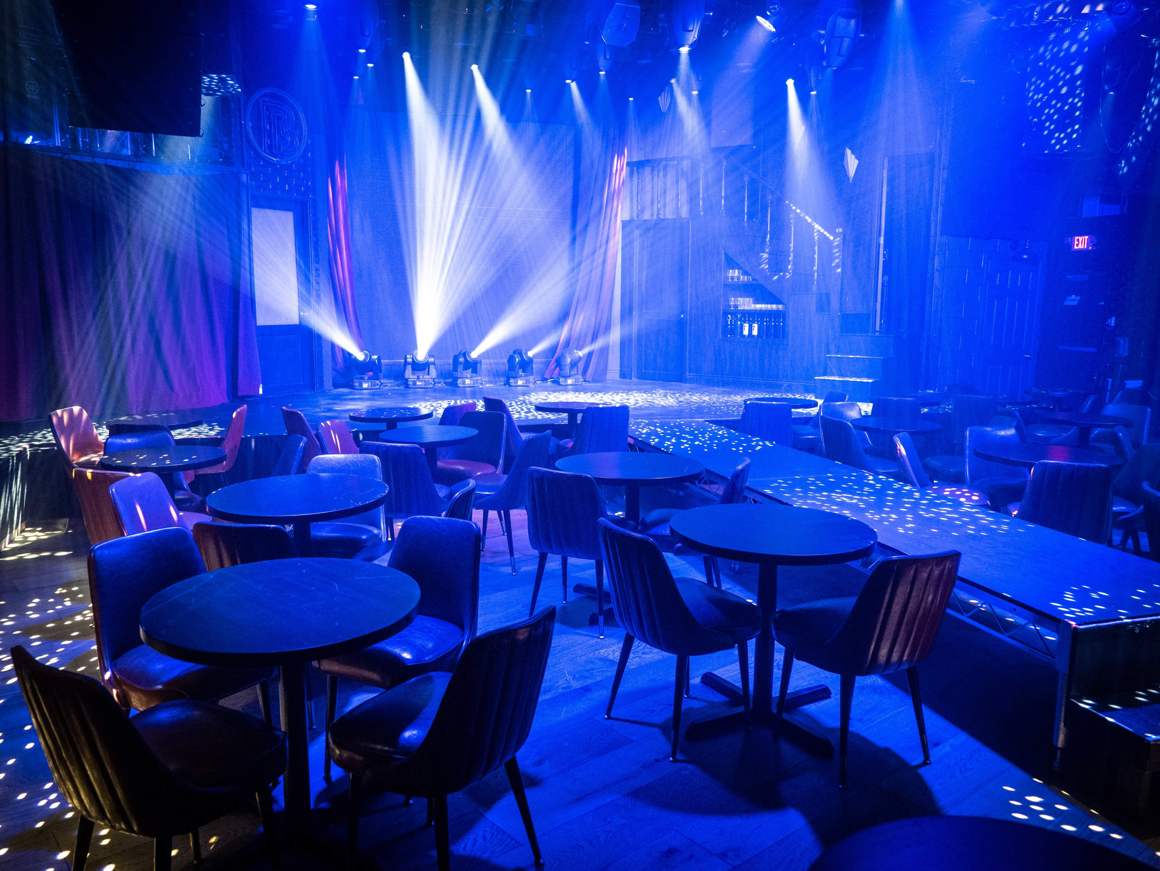 A dimly lit performance venue features circular tables and chairs with stage lights casting beams across the floor.