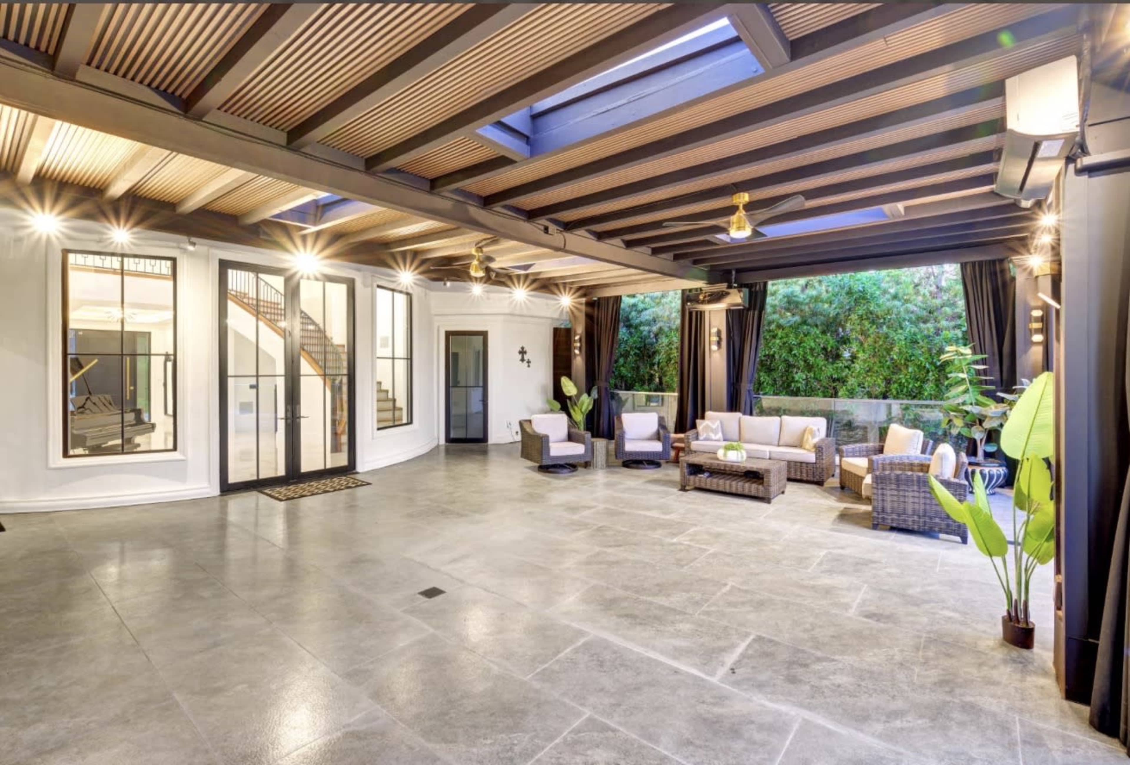 The image shows a spacious open living area with modern furniture, surrounded by large windows and lush greenery visible outside.