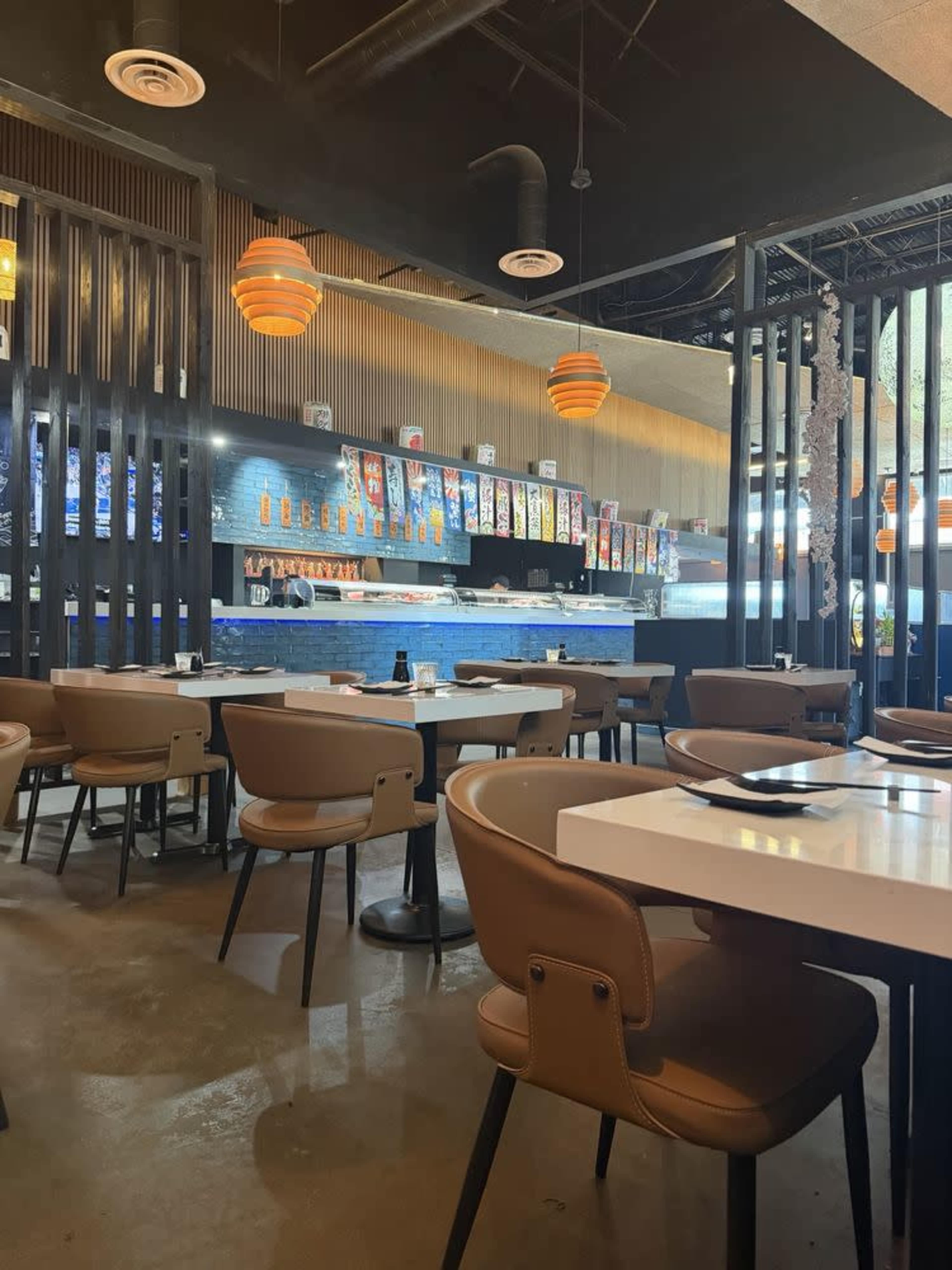 The image shows an interior view of a modern restaurant with tables and seating arranged around a bar area, featuring bright pendant lights and a display of menus on the wall.