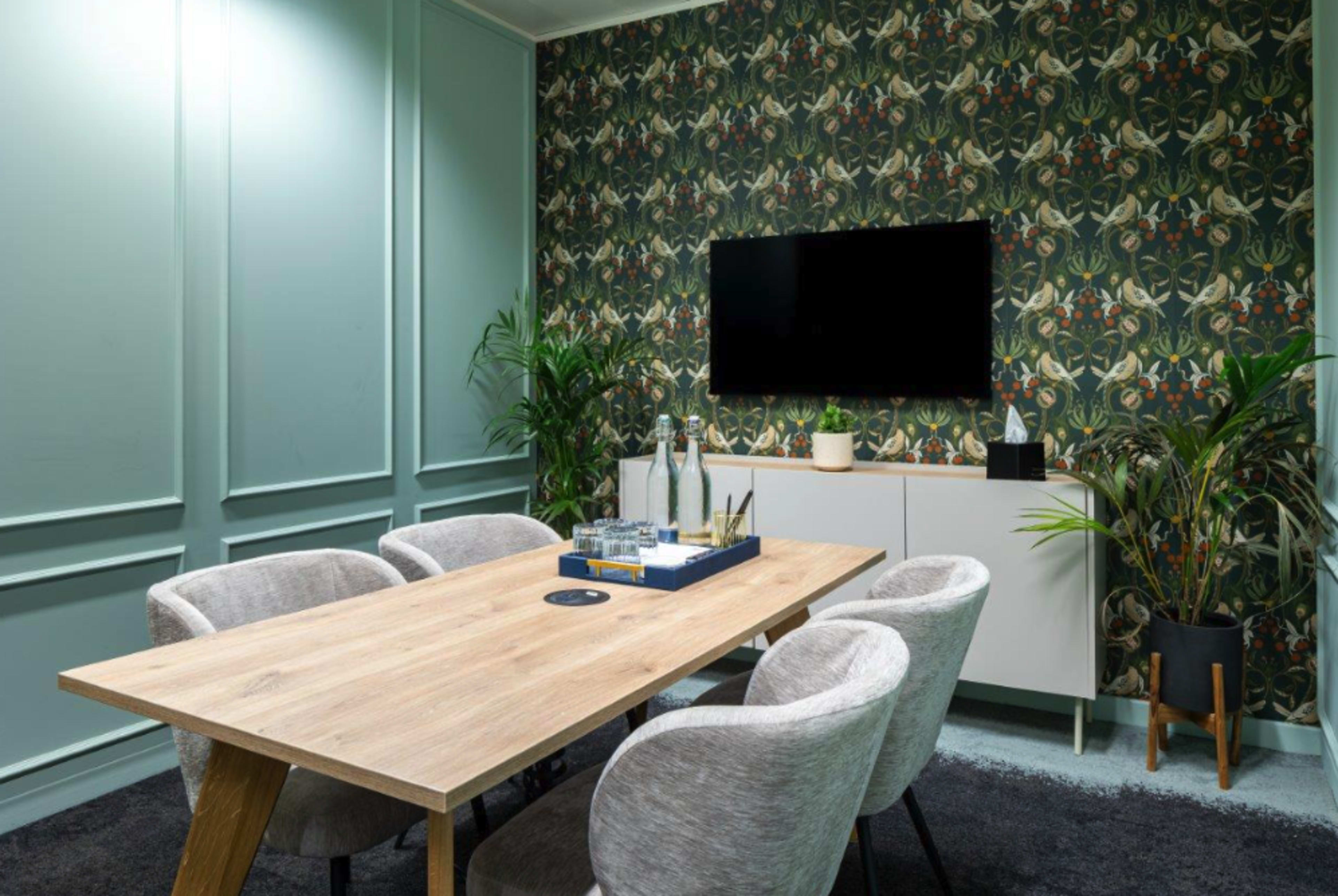 The image shows a modern conference room featuring a wooden table surrounded by four upholstered chairs, with a patterned wallpaper and a television mounted on the wall.