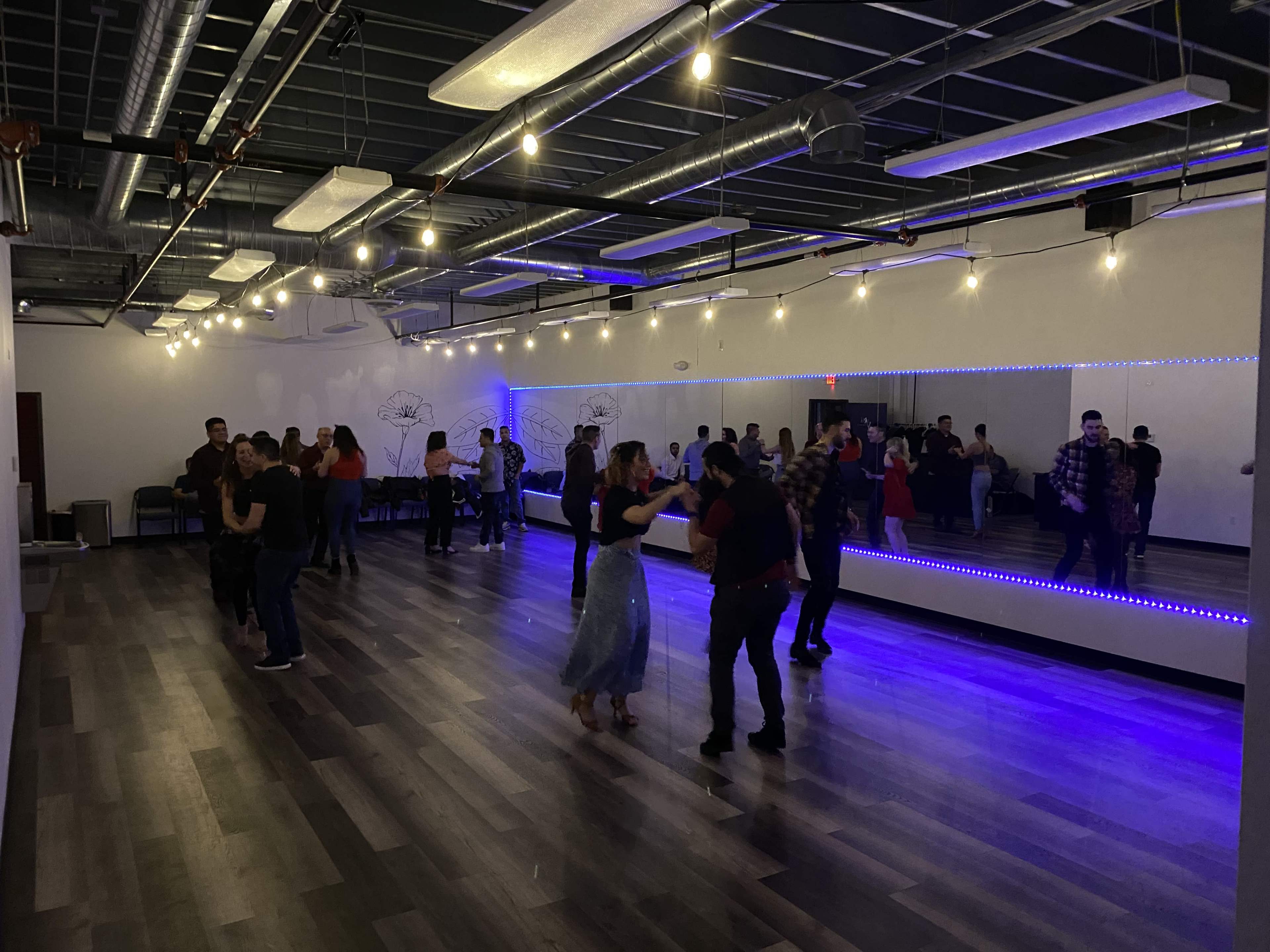 A group of people dance on a wooden floor in a well-lit studio with mirrors along one wall.