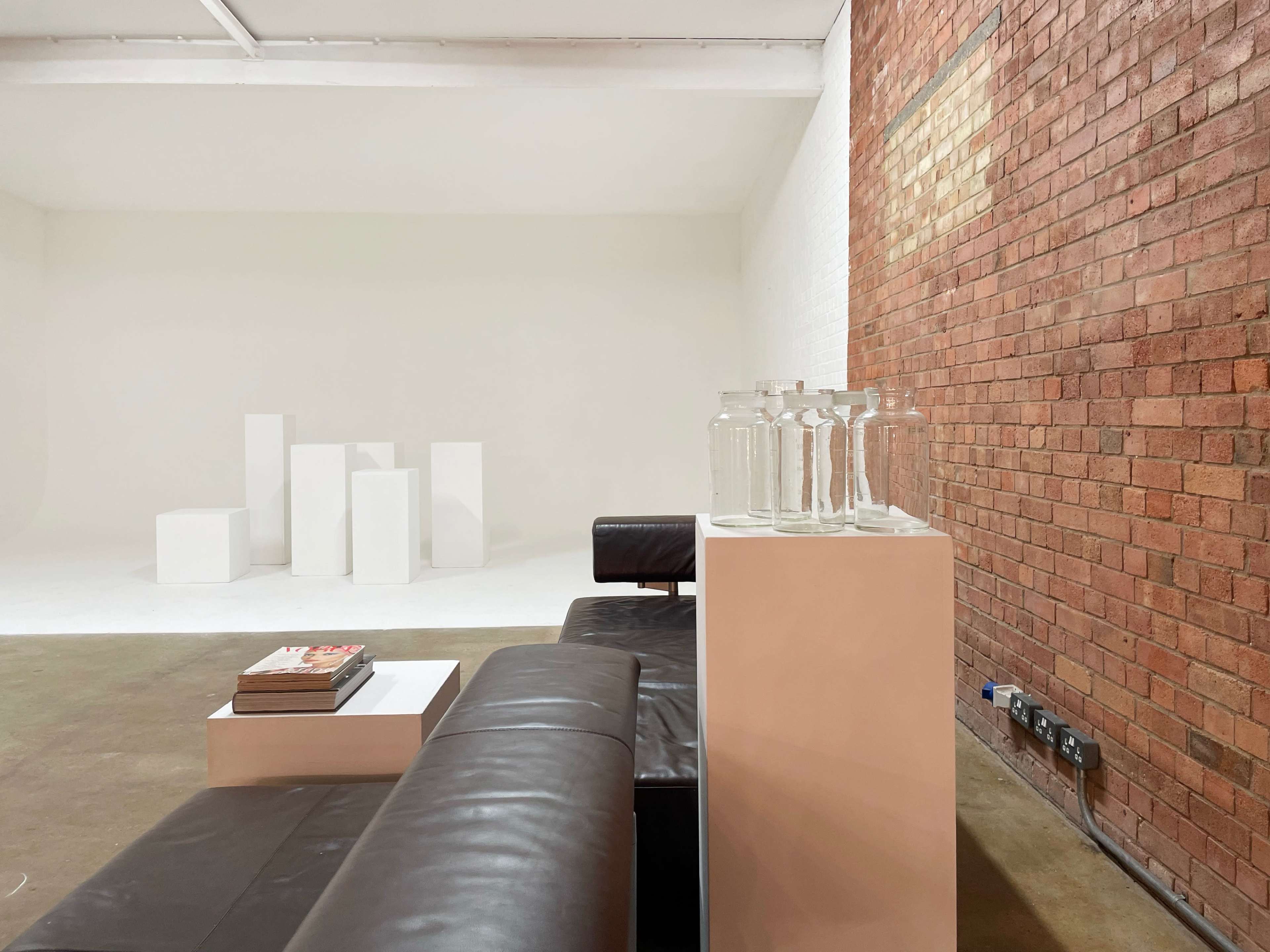 A minimalist interior with a black leather sofa, a white coffee table adorned with magazines, and several transparent glass jars, all set against a backdrop of a textured brick wall and white pedestals.