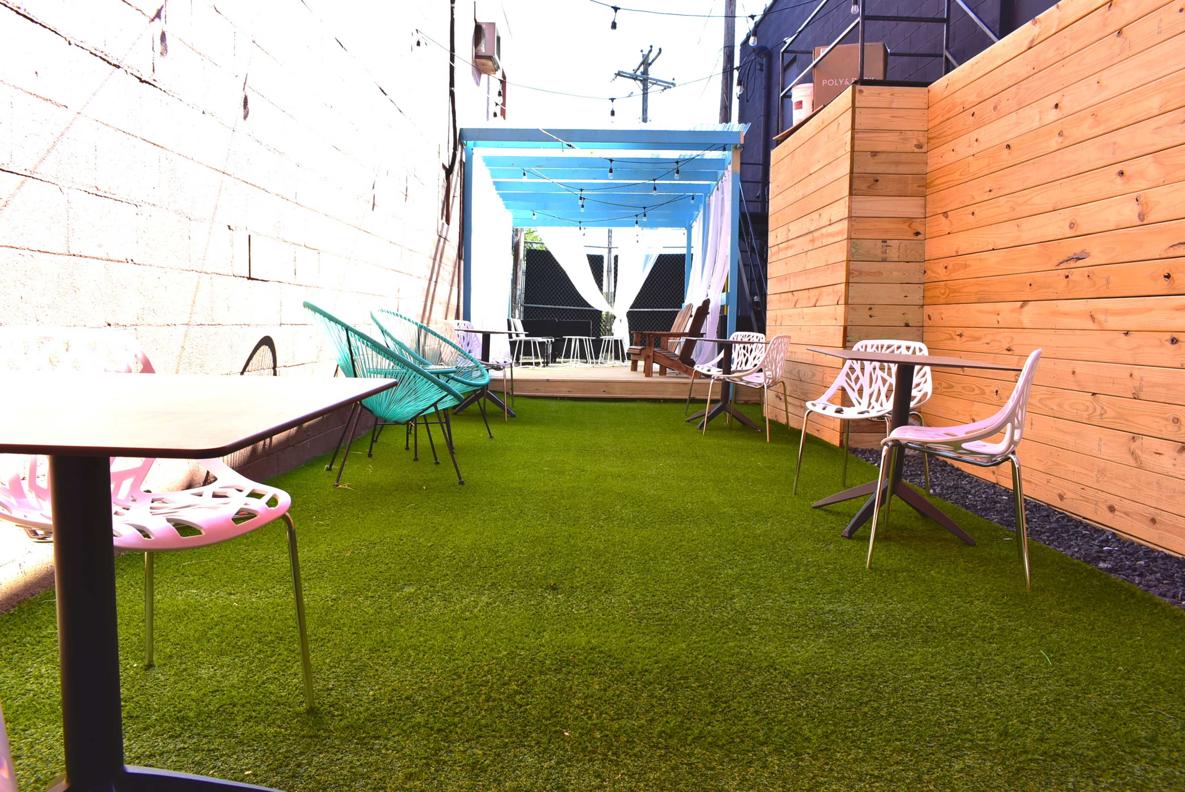 A narrow outdoor space with artificial grass, featuring tables and chairs along the sides and a blue canopy overhead.