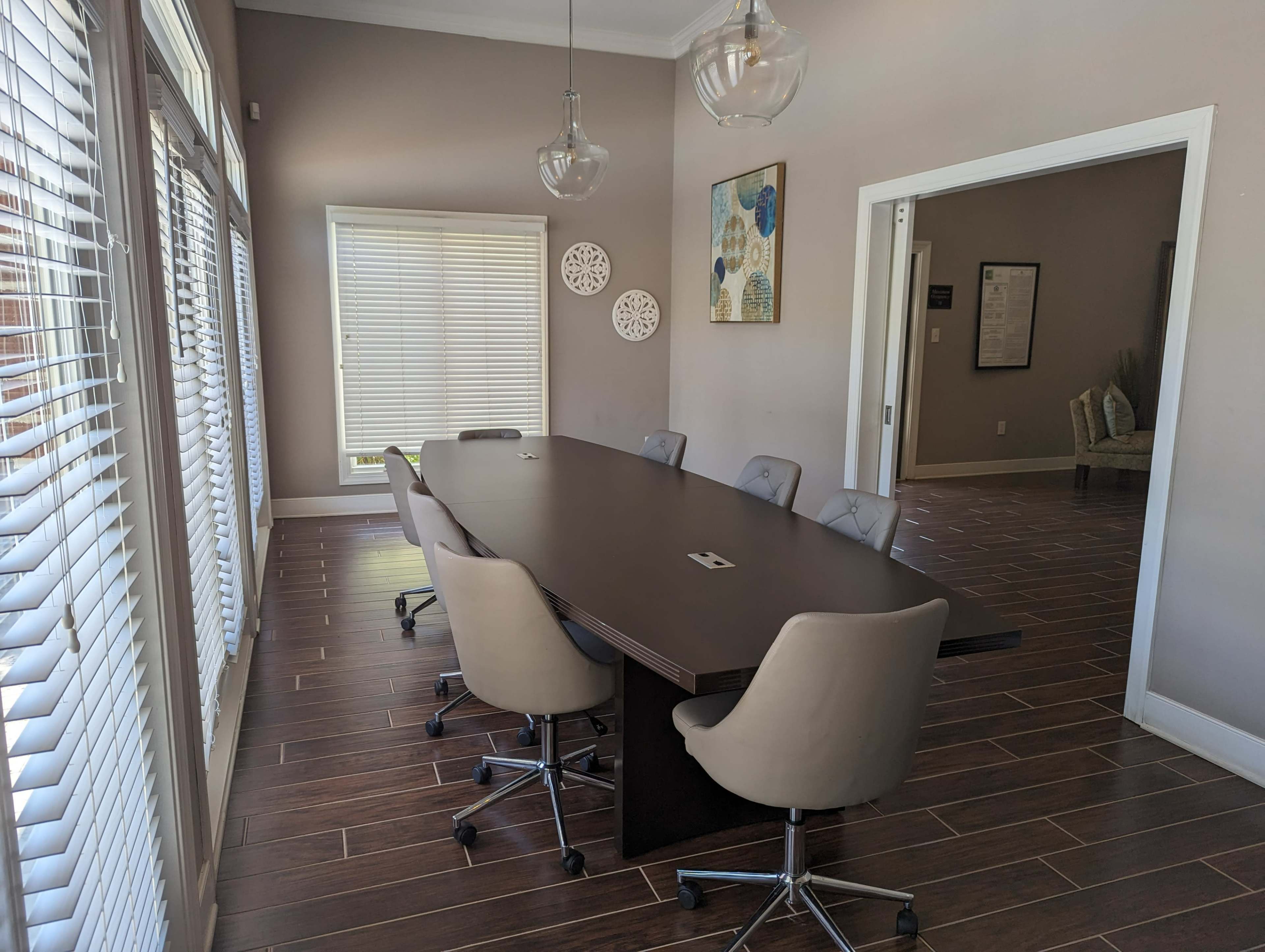 A conference room features a long dark table surrounded by six light-colored swivel chairs, with large windows allowing natural light to illuminate the space.