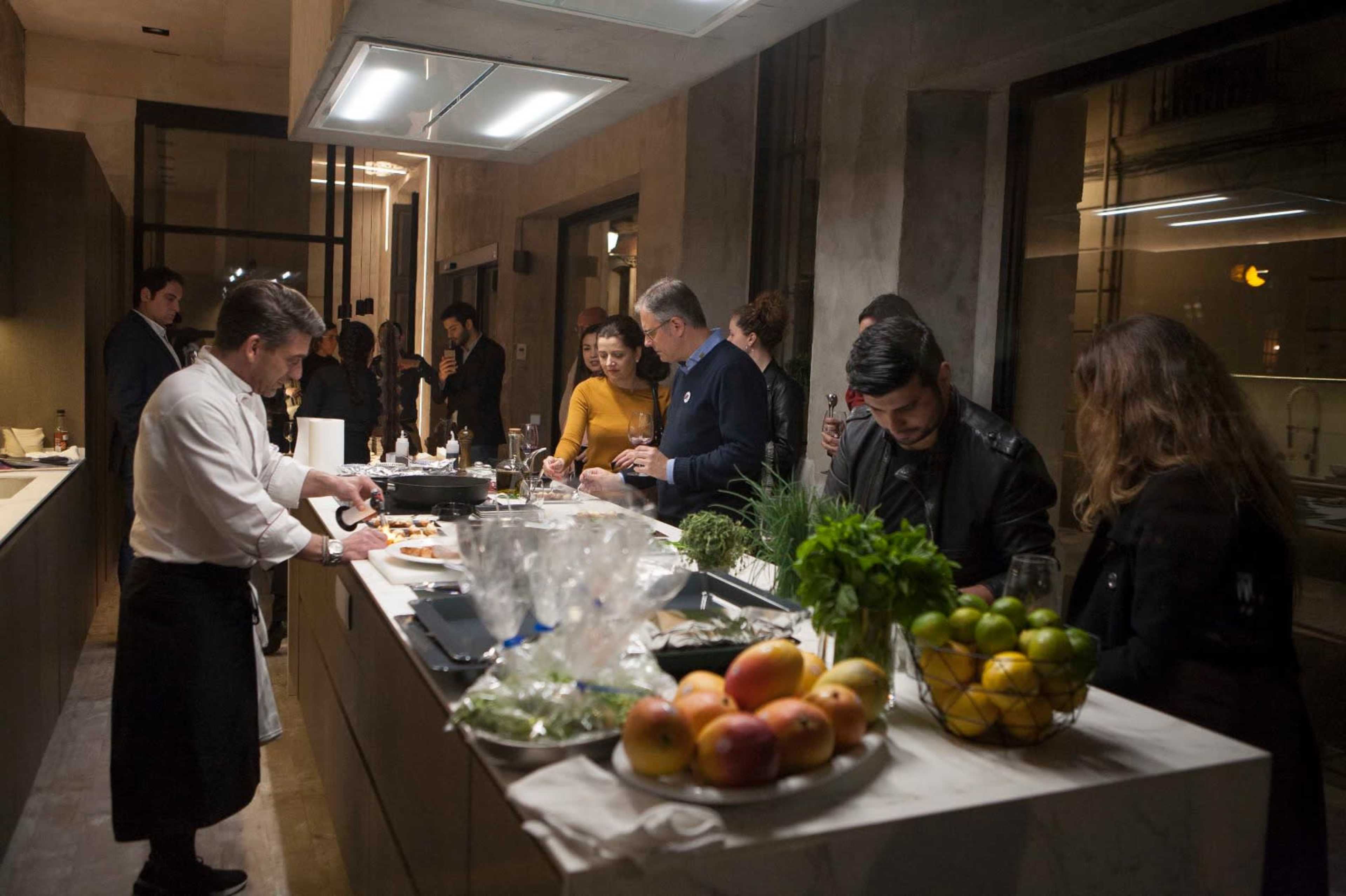 A group of people socializes in a modern kitchen while a chef prepares food on the counter, surrounded by fresh ingredients and fruits.