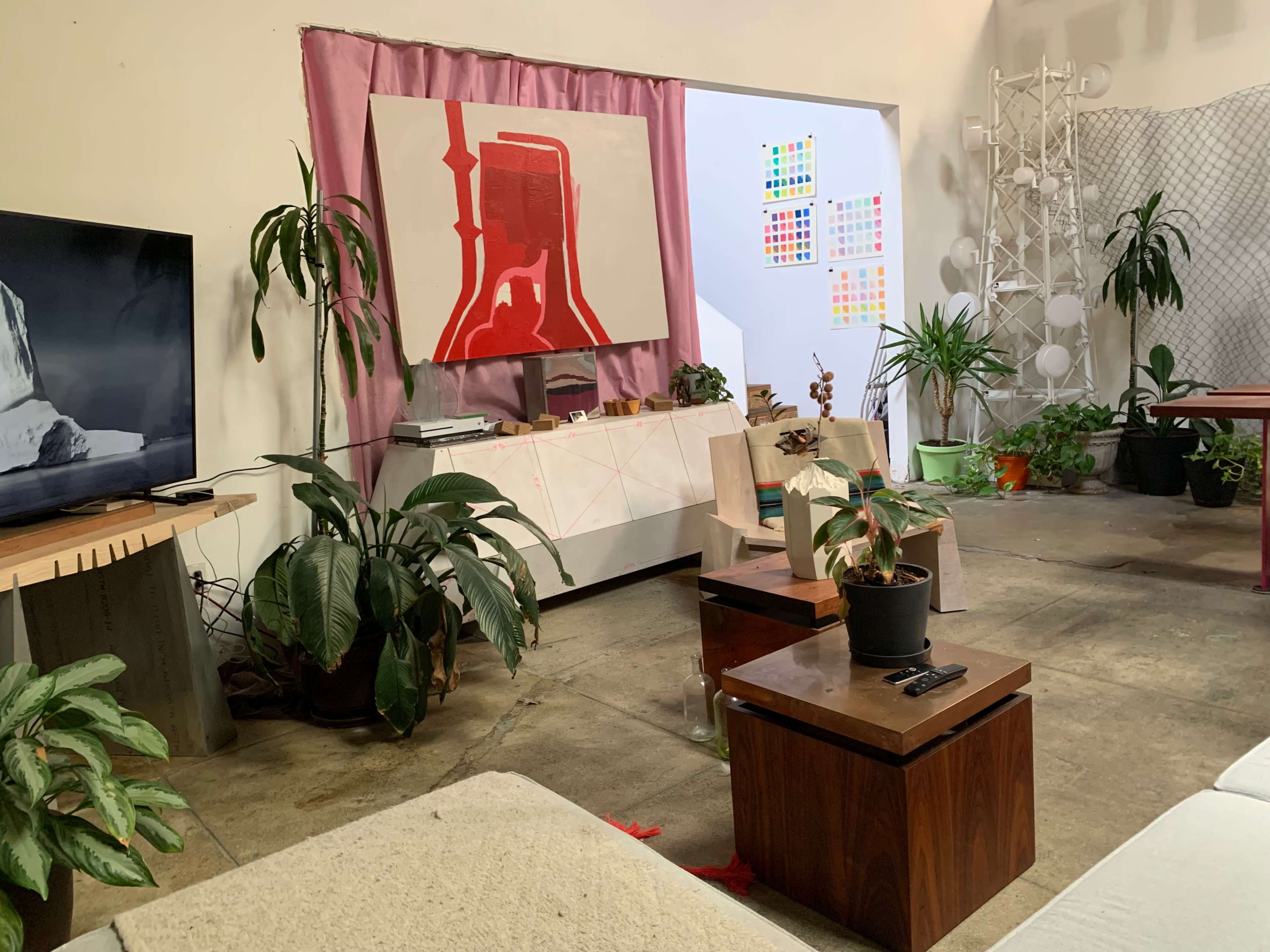 The image shows a creatively arranged indoor space with plants, a television, and artwork on display, along with seating areas and a colorful wall installation.