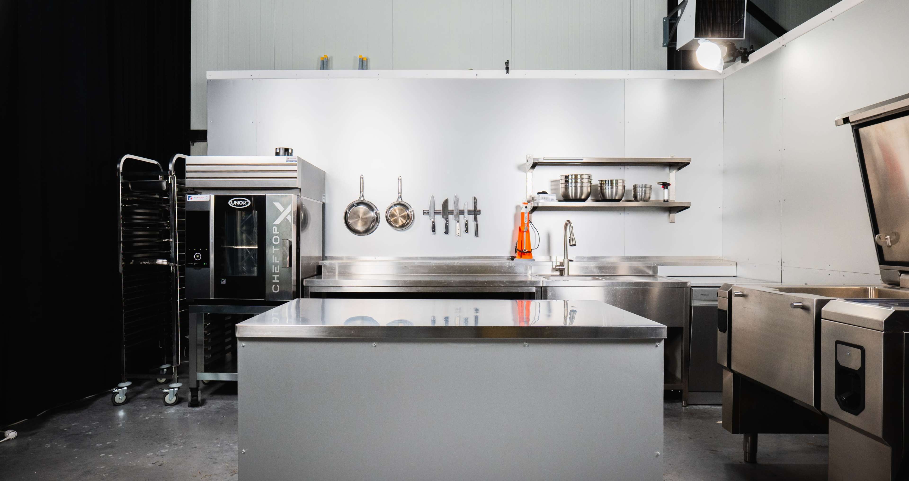 The image shows a modern kitchen setup featuring stainless steel counters, a baking oven on the left, and various kitchen utensils hanging on the wall.