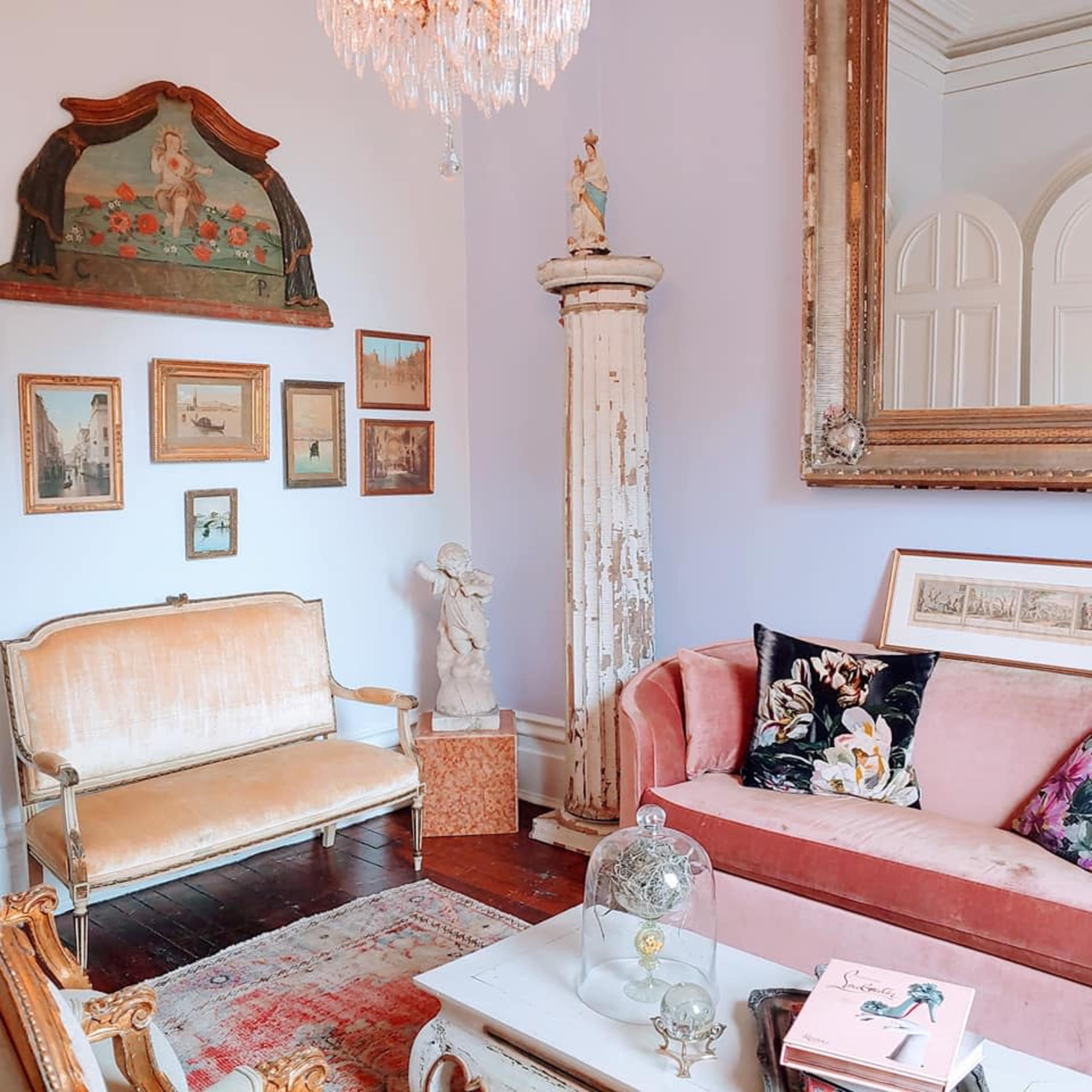 The image shows a tastefully decorated living room with a vintage sofa, ornate mirror, framed artwork, and a chandelier, all set against a pastel wall.