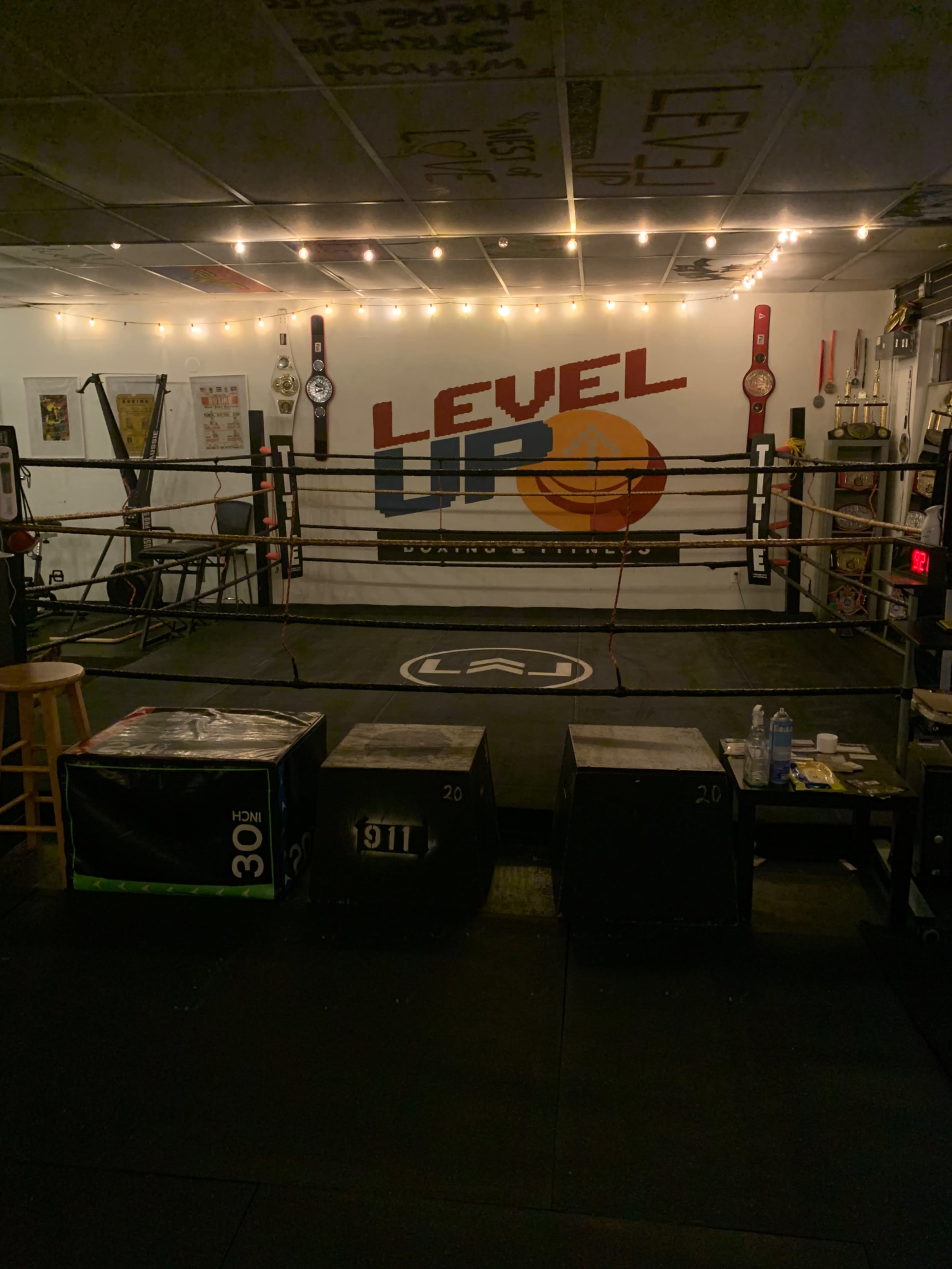 A boxing ring in a dimly lit training facility, surrounded by exercise equipment and decorated with championship belts on the walls.
