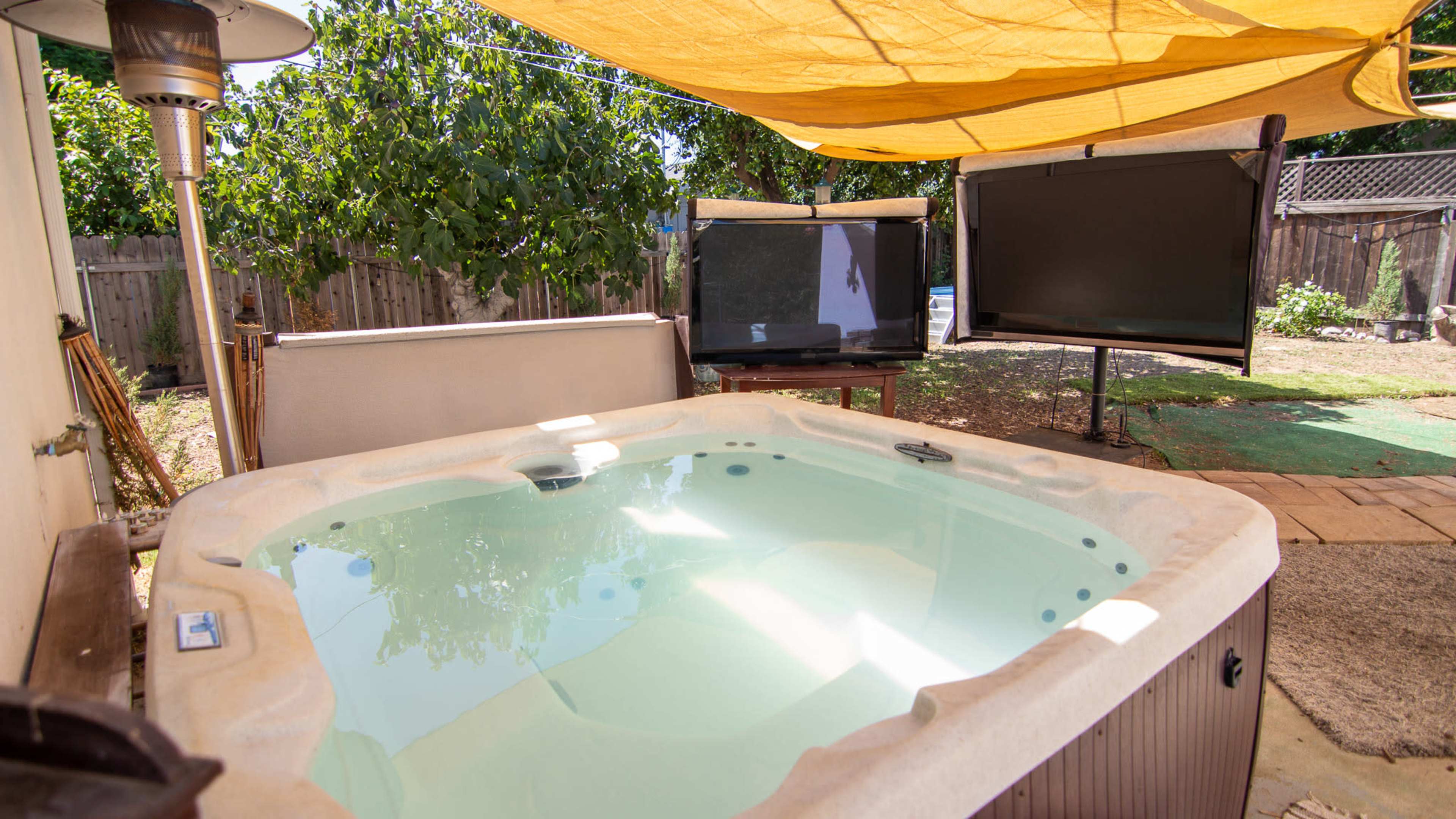 A hot tub under a yellow canopy in a backyard, with two large screens positioned nearby.
