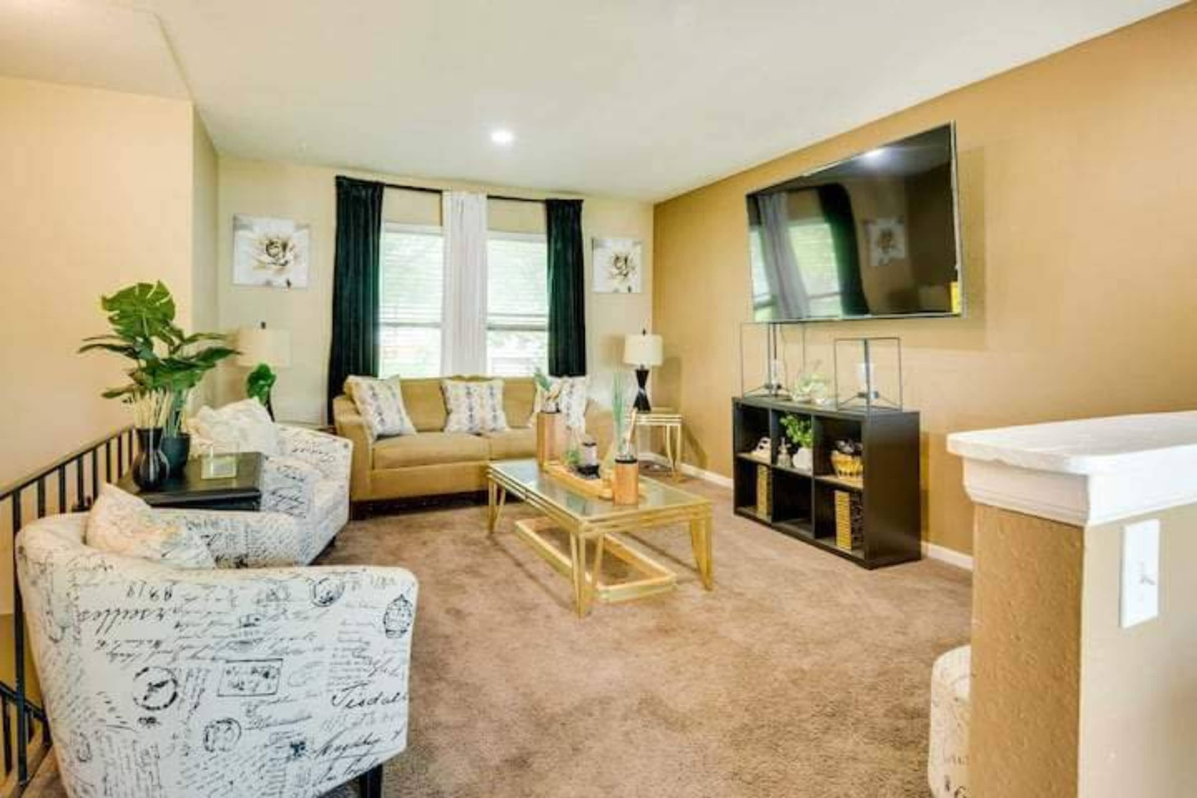 A modern living room featuring a tan sofa, two patterned chairs, a glass coffee table, and a large wall-mounted television.