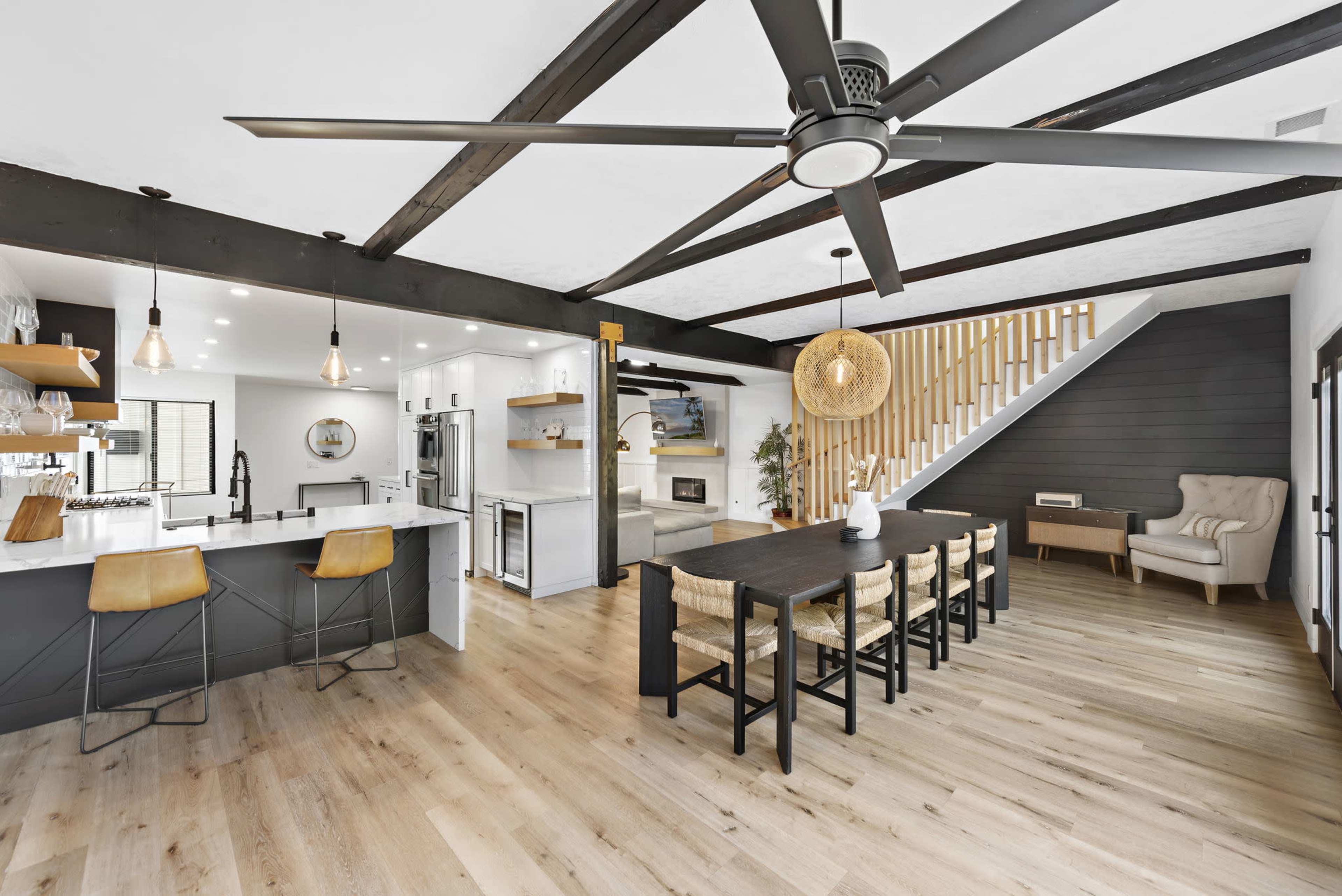 The image shows a modern open-concept kitchen and dining area with a large wooden table, bar seating, and a staircase in the background.