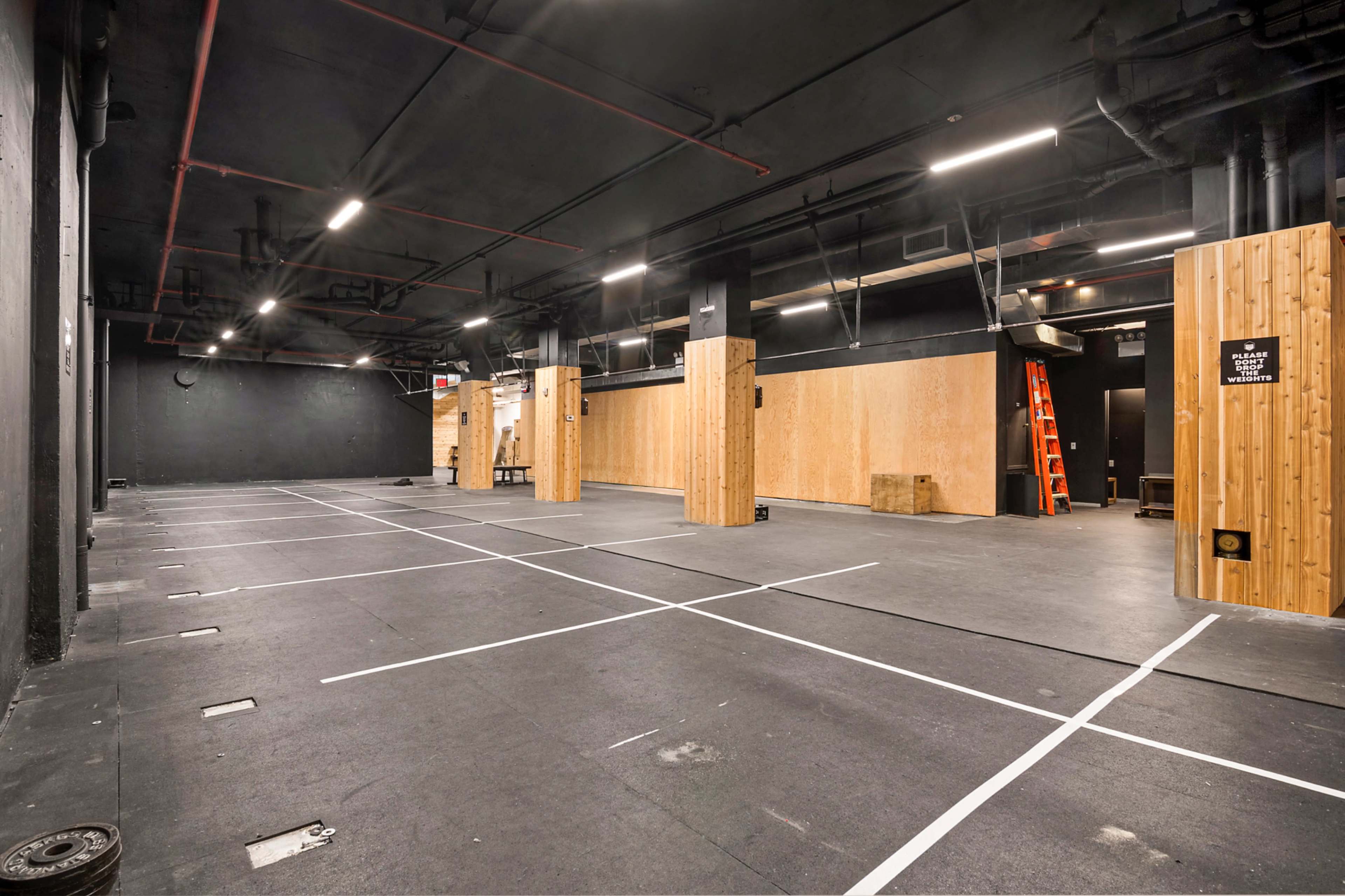 The image shows an empty gym space with black walls, wooden columns, and marked white workout zones on the floor.