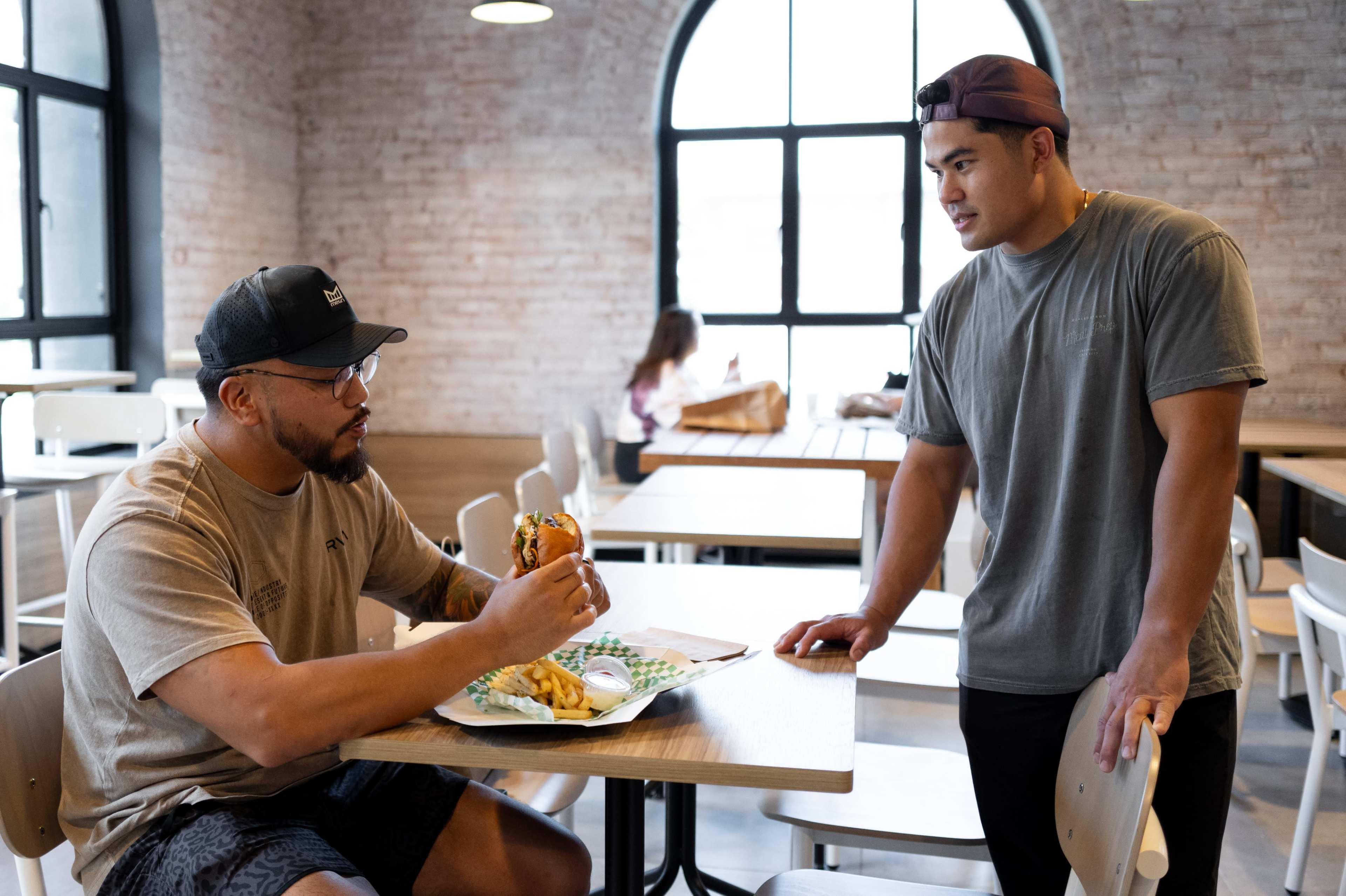 Two men are in a casual dining setting, with one man holding a burger and fries while the other stands nearby engaged in conversation.