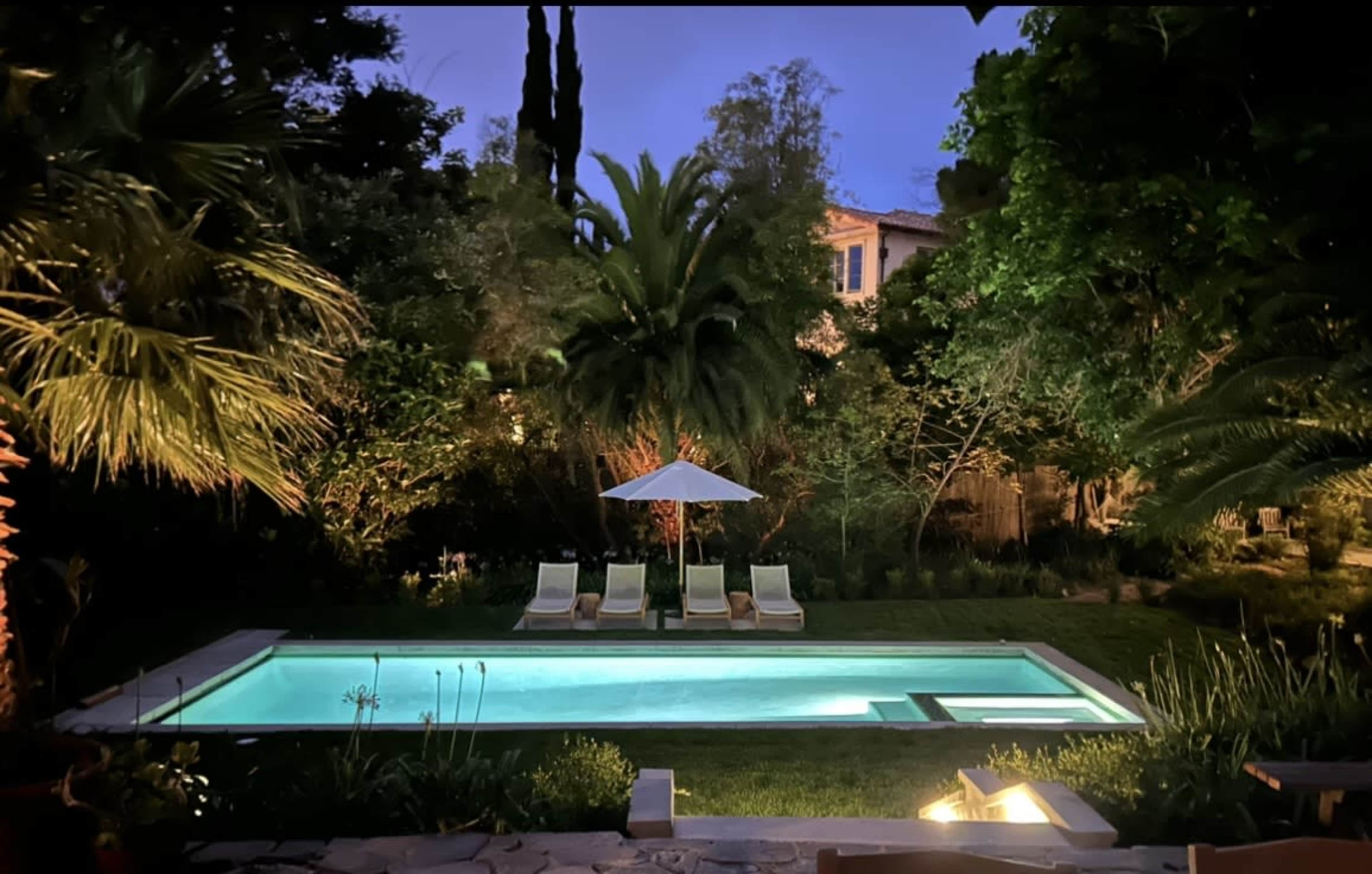 A well-lit swimming pool surrounded by lush greenery and lounge chairs, with an umbrella in the background under a twilight sky.