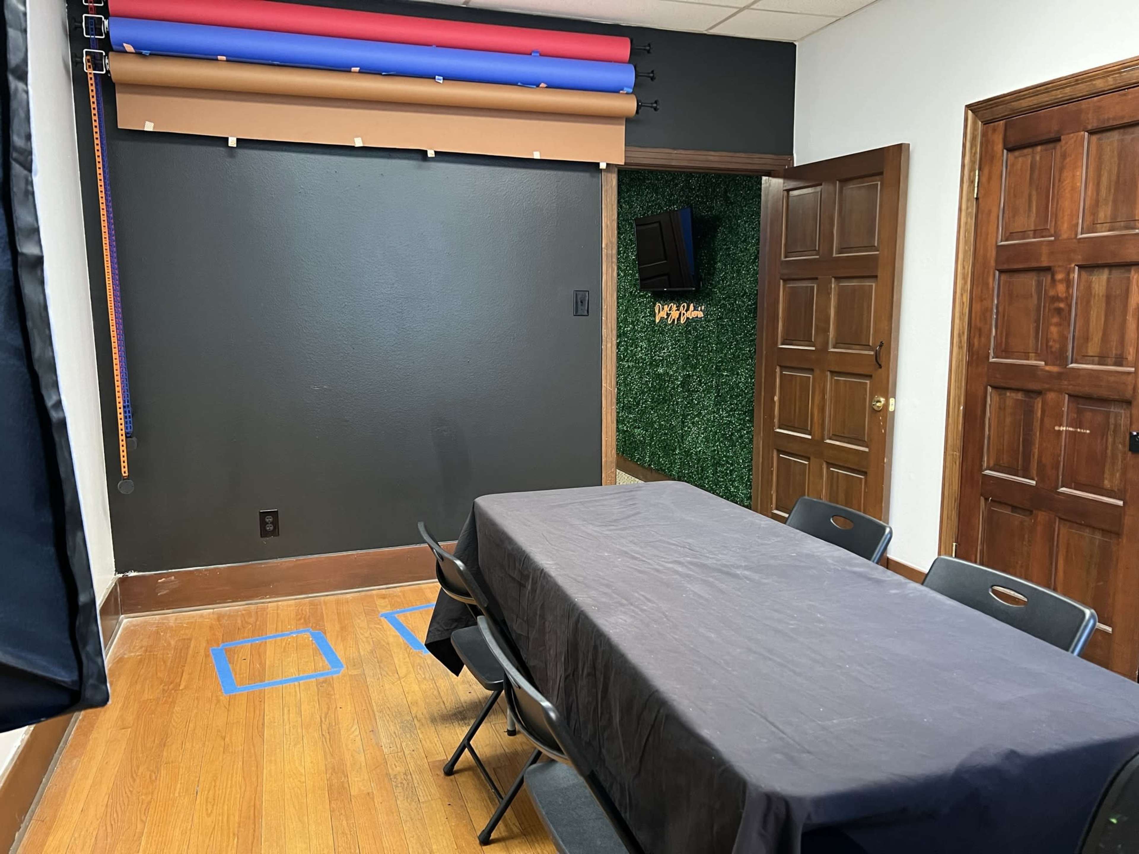 A room with a black wall, a table covered with a dark tablecloth, and chairs arranged around it, with a green accent wall and a small TV mounted nearby.