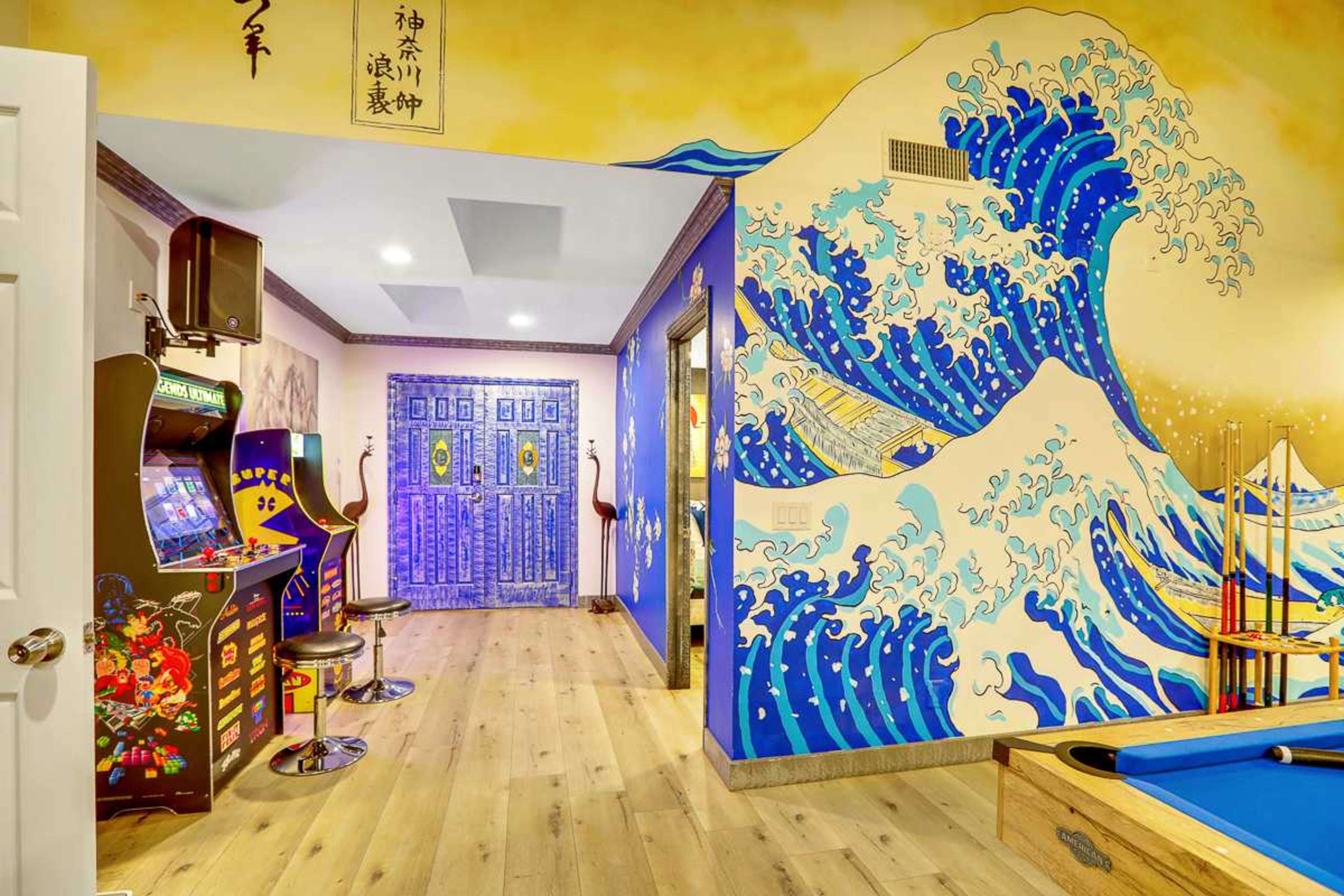 A brightly colored game room with a large mural of waves on one wall, a door designed with intricate patterns, and arcade games positioned beside a pool table.