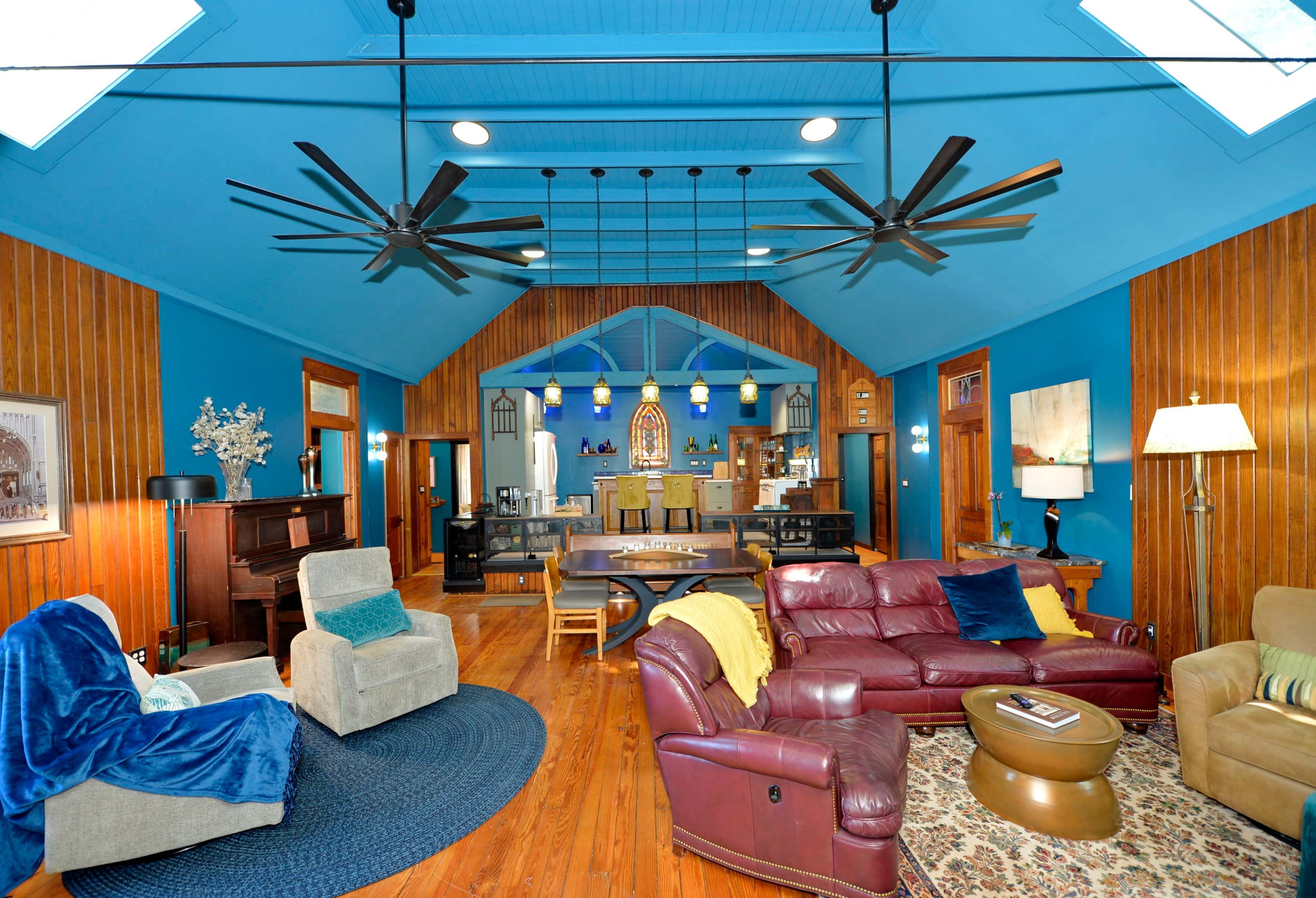 The image shows a spacious living area with blue walls, wooden accents, and large ceiling fans, featuring various seating arrangements and a piano.
