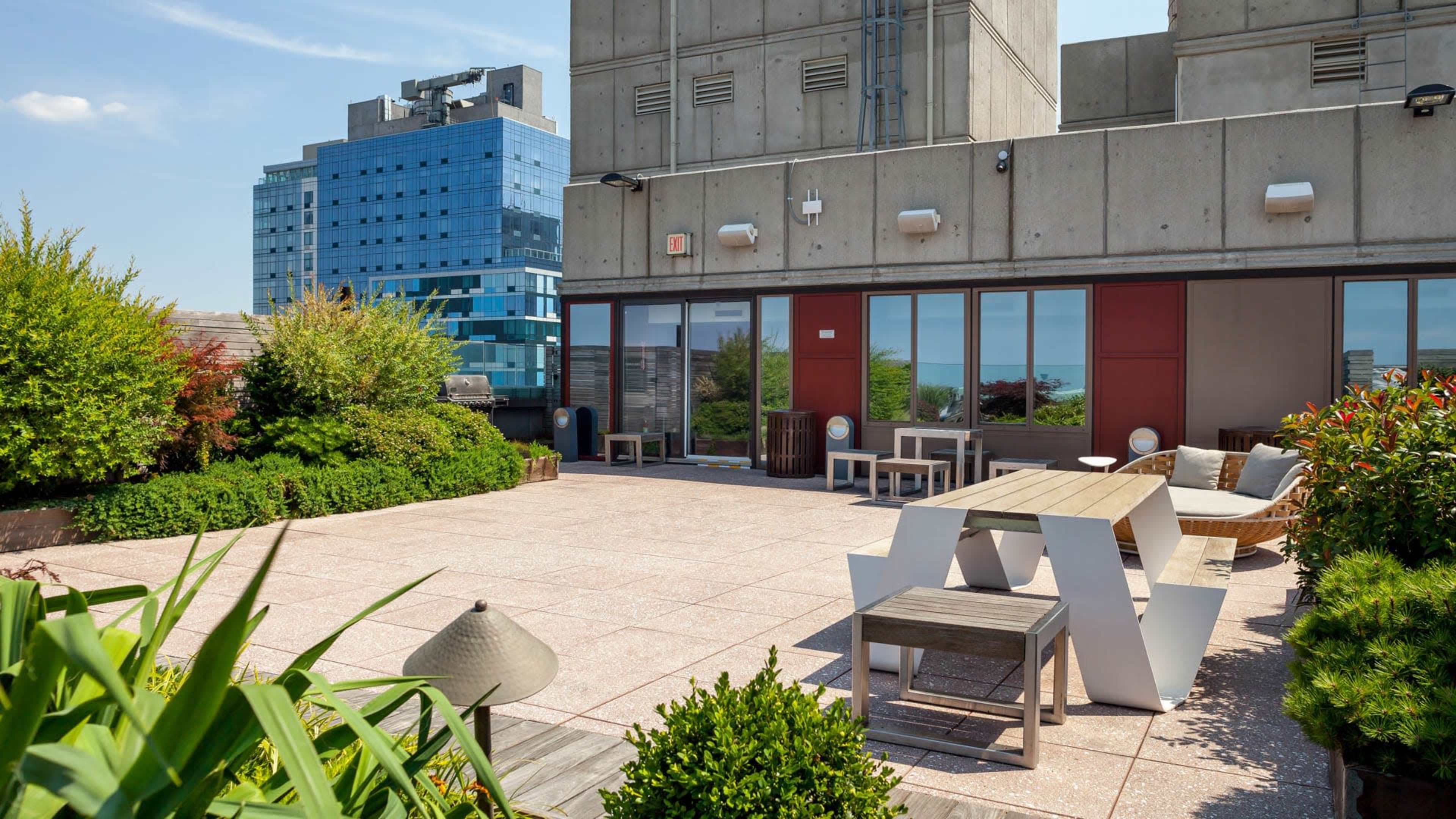 A rooftop terrace with modern furniture, plants, and a view of nearby buildings and the sky.