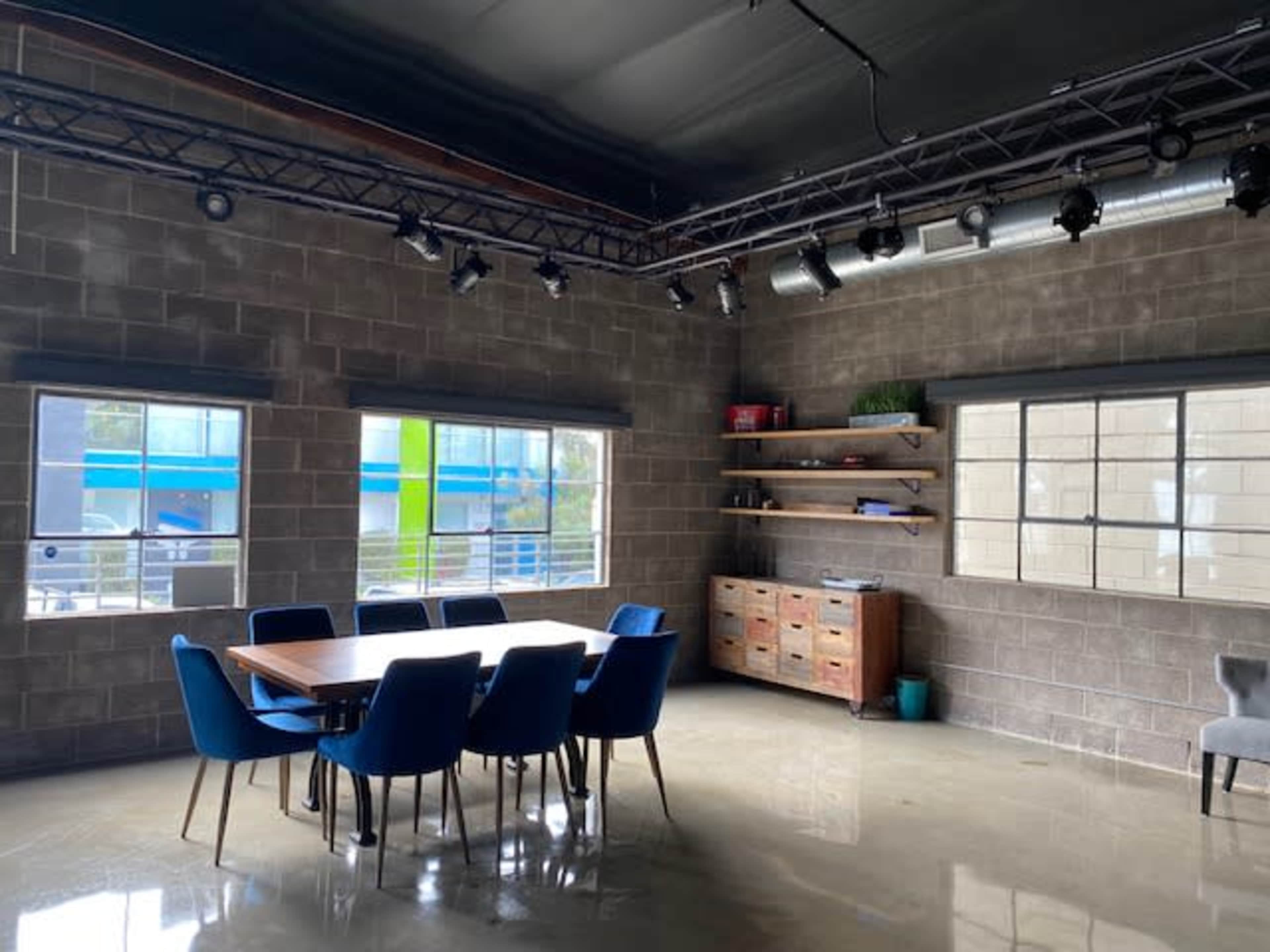 A conference room features a large wooden table surrounded by blue chairs, with windows allowing natural light to enter and industrial-style walls.