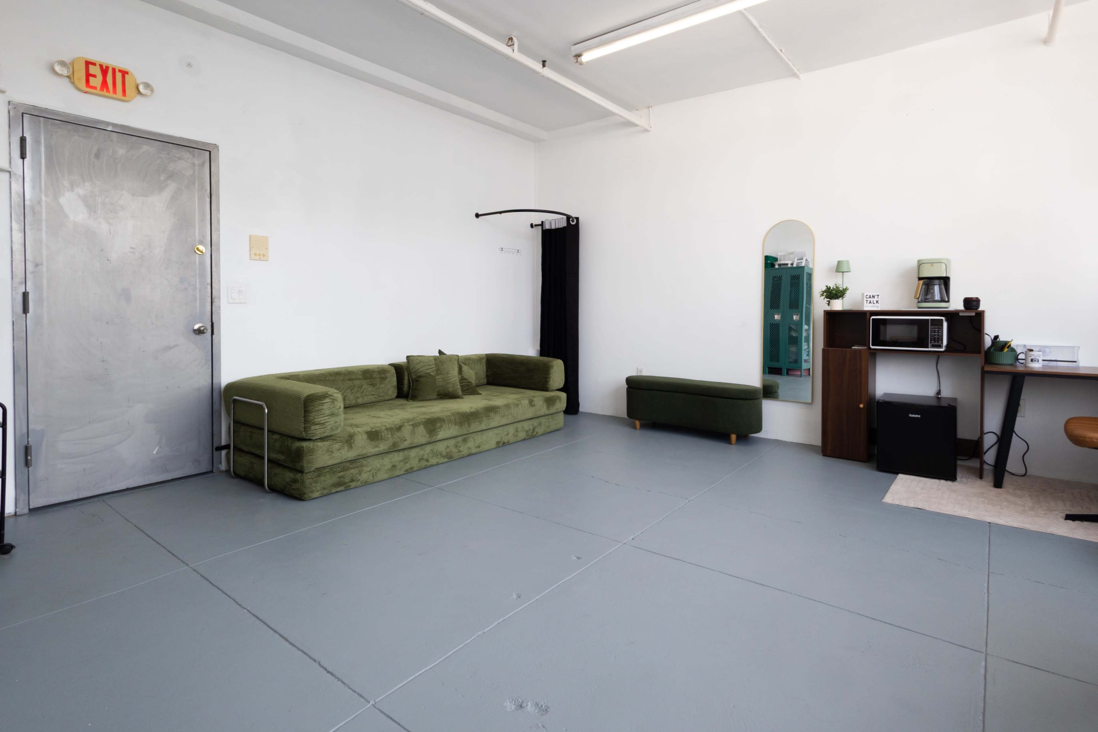 The image shows a minimalist room with a green sofa, a small matching ottoman, a mirror on the wall, a microwave, and a desk in one corner.