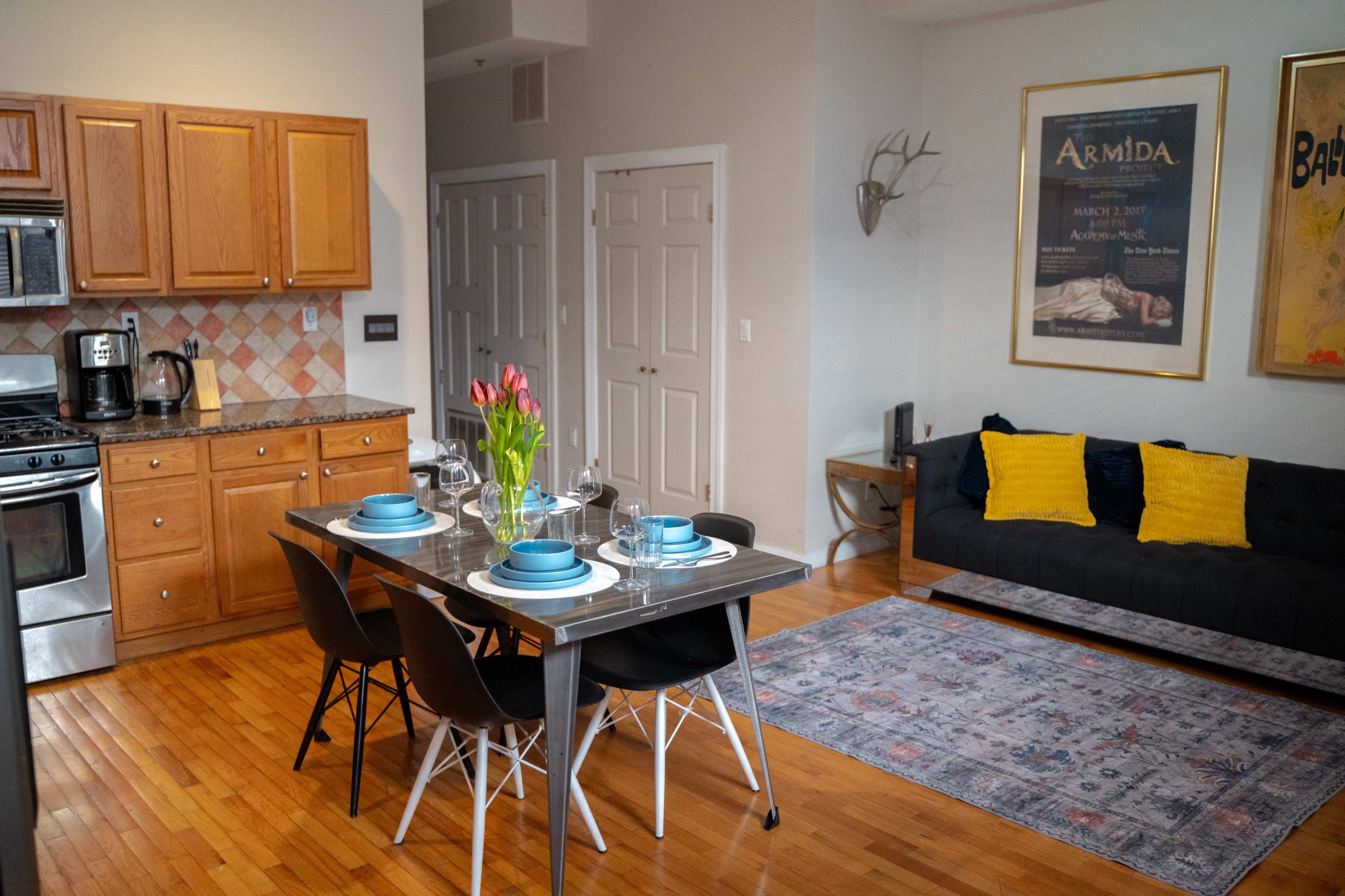 The image shows a dining area with a table set for four, a kitchen with wooden cabinets, and a cozy living room featuring a black couch and decorative artwork on the walls.