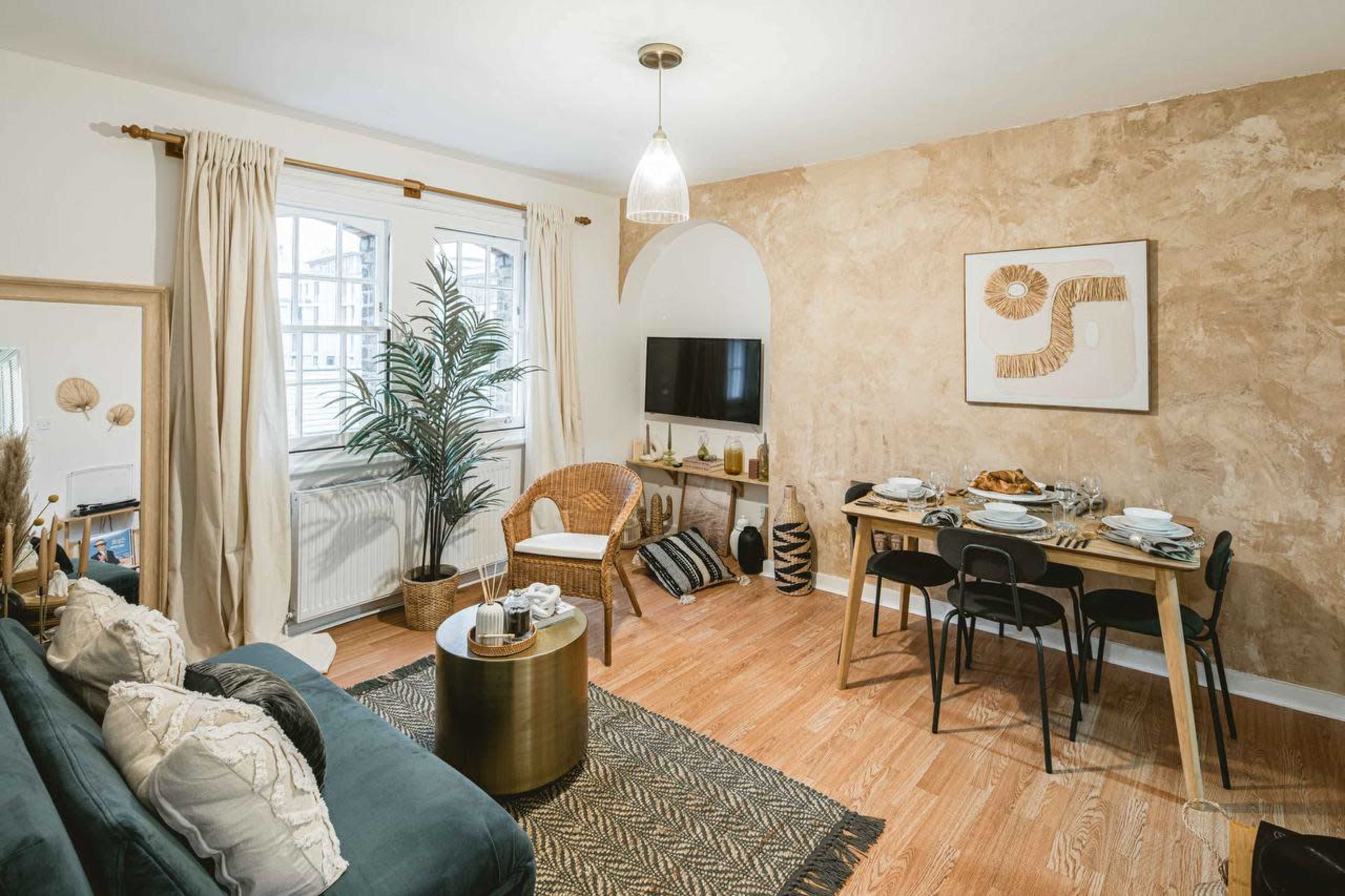 The image shows a cozy living room with a green sofa, a round coffee table, a dining area with wooden chairs, and a decorative wall featuring a piece of art.