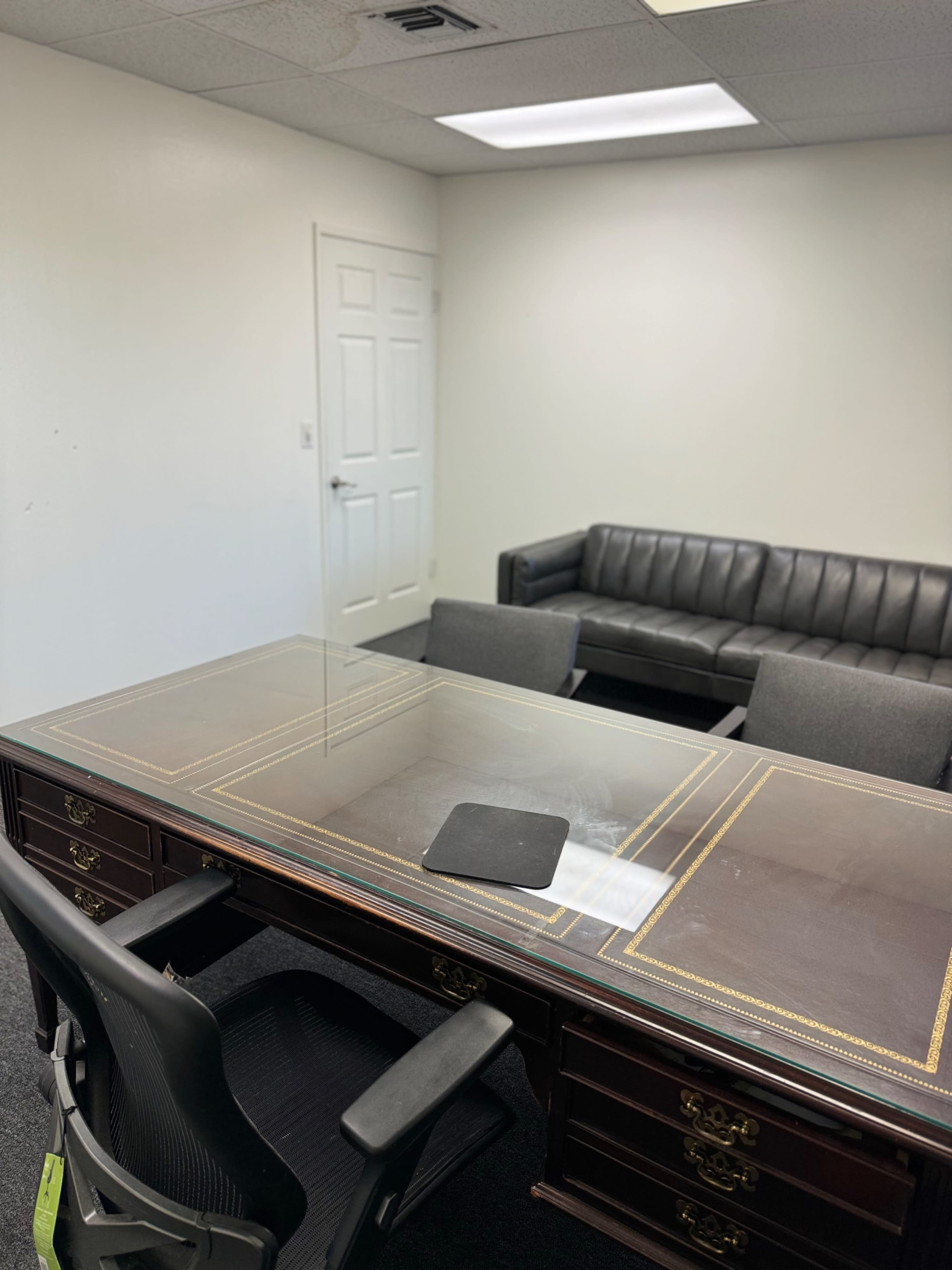 The image shows a simple office space featuring a wooden desk with a glass top, a rolling chair, and a gray couch along with two gray chairs.